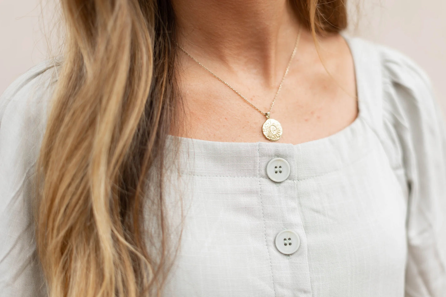 Close-up of a person wearing a light gray Lydia Embroidered Midi Dress from Dogwood Cloth, featuring flutter sleeves and complemented by a gold necklace with a round pendant.