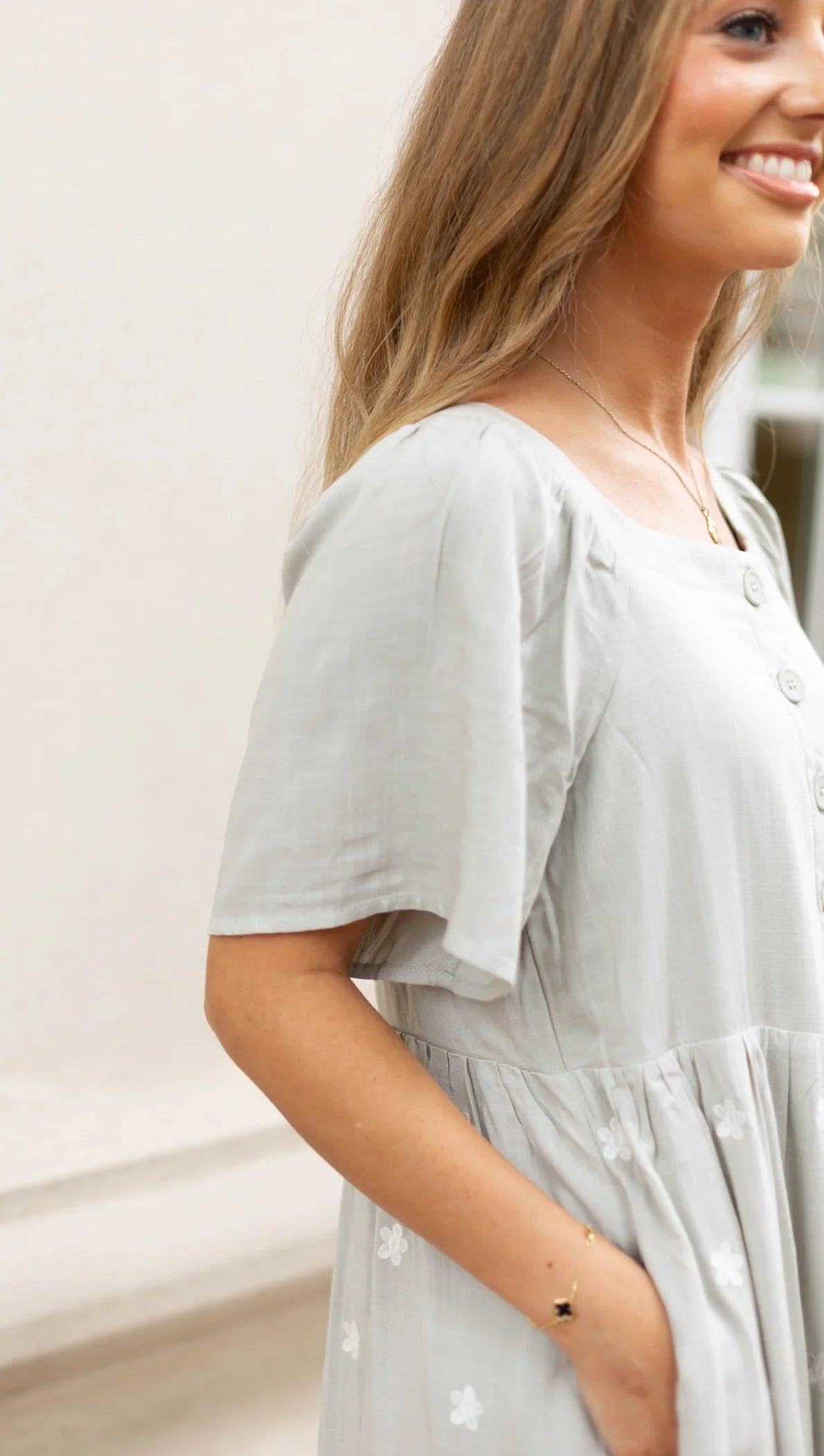 A person in a light gray Lydia Embroidered Midi Dress by Dogwood Cloth, featuring pockets and flutter sleeves, facing sideways against a neutral background.