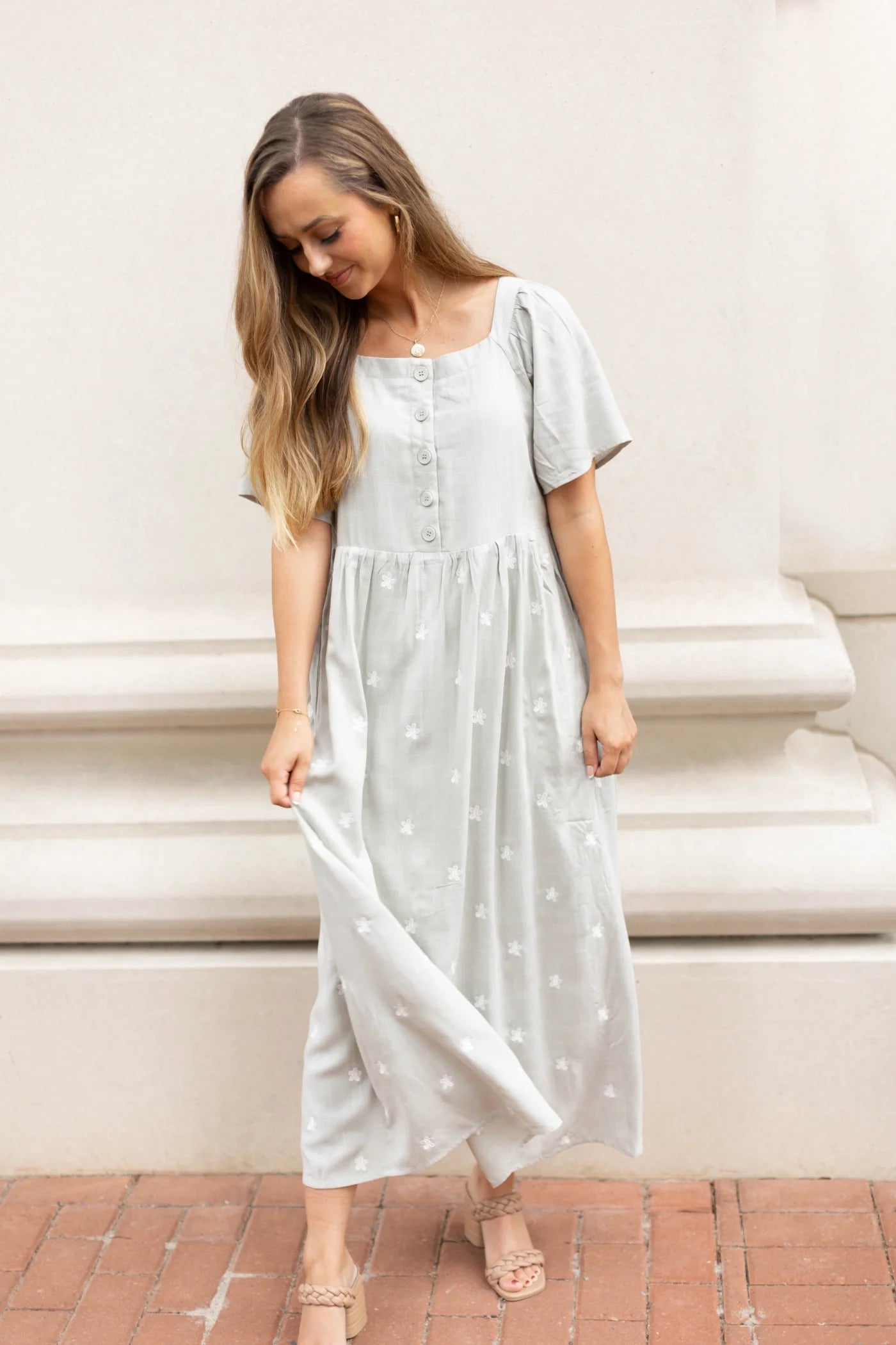 A woman wearing the Dogwood Cloth Lydia Embroidered Midi Dress stands on a brick surface. She looks down while gently holding the hem of her elegant dress with one hand.