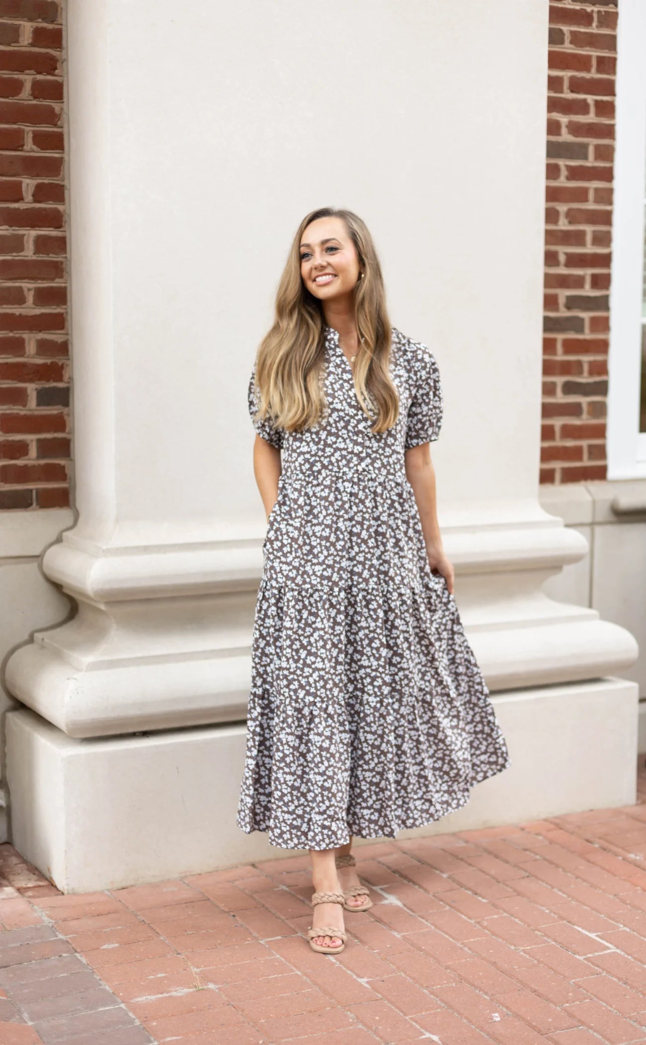 A woman stands outdoors wearing a playful Ava Tiered Midi by Dogwood Cloth, featuring vibrant floral patterns and side pockets with short sleeves. She is smiling and has long hair, set against the backdrop of a white architectural structure and a brick wall.