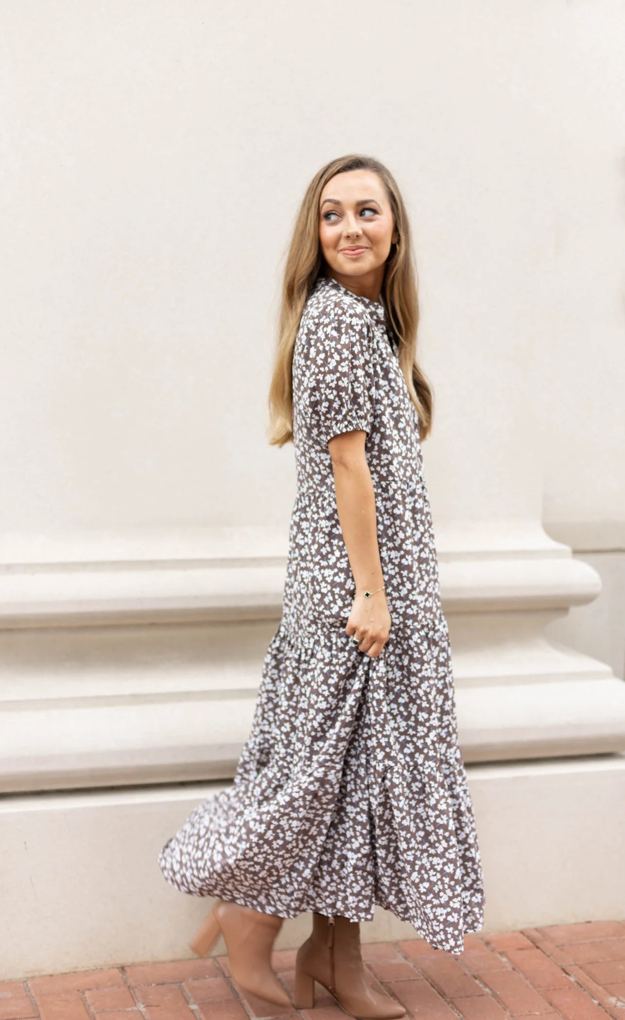 A woman in a long, floral Ava Tiered Midi from Dogwood Cloth stands on a brick pavement, exuding playful elegance as she glances to the side. Her flowing locks beautifully complement the dress's design, while subtle side pockets provide both style and function near the pristine white wall.