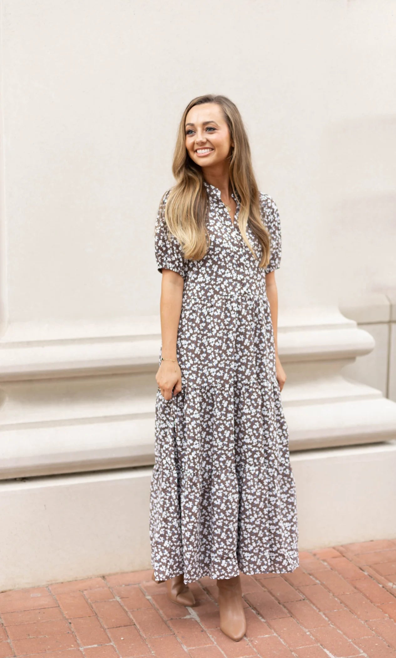 A person wearing a floral Ava Tiered Midi dress by Dogwood Cloth stands smiling on a brick pathway, exuding playful elegance against a neutral-colored wall, with the subtle charm of side pockets adding to the ensemble.