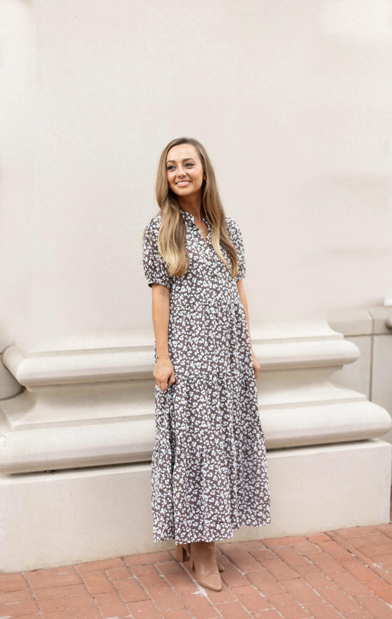 A person wearing the Ava Tiered Midi by Dogwood Cloth stands on a brick sidewalk against a cream-colored wall, exuding playful elegance with their smile.