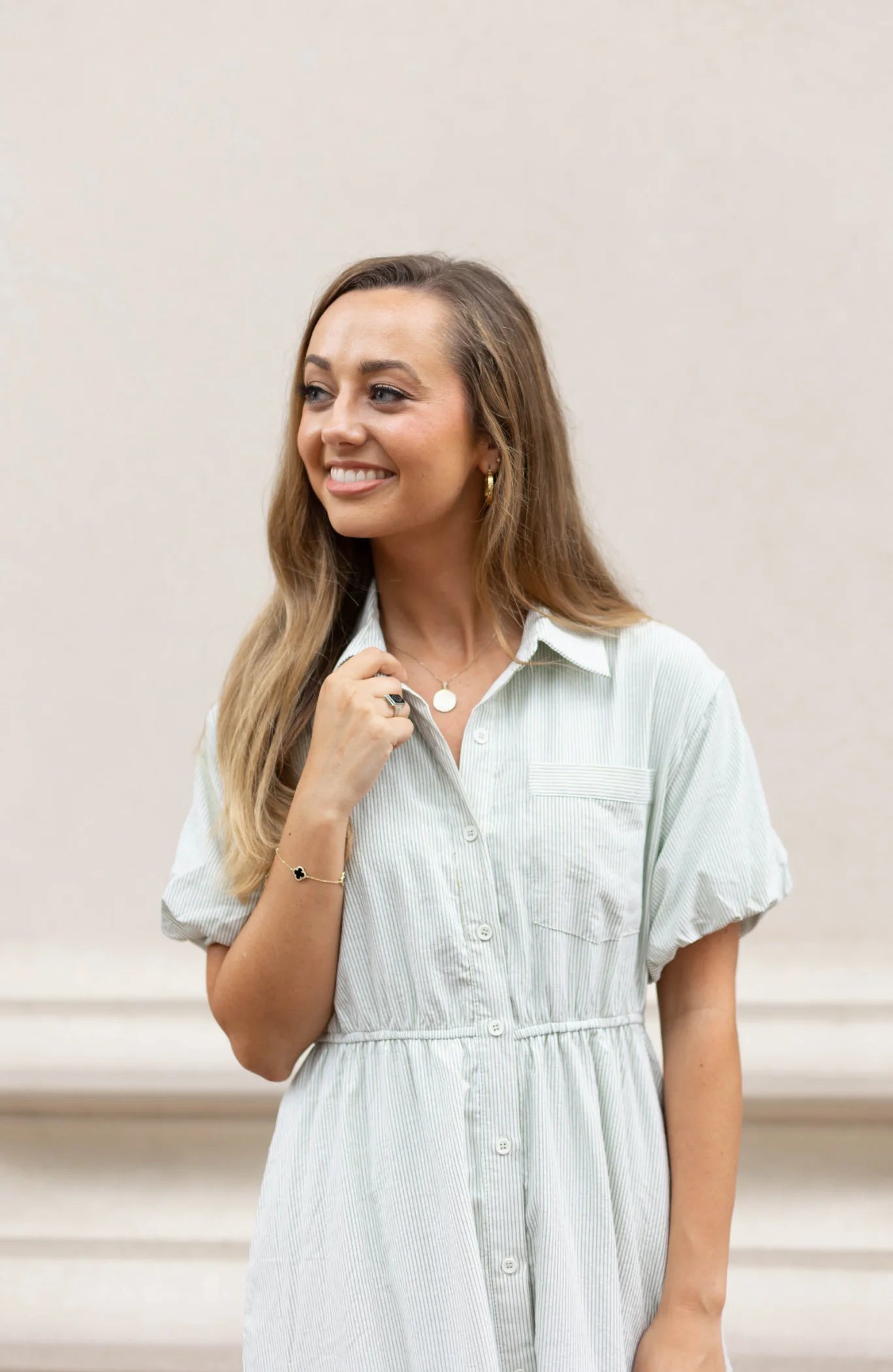 A smiling woman in a light-colored Carson Pin-Stripe Midi Dress by Dogwood Cloth, featuring an elastic waistband, stands against a plain backdrop, looking to the side.