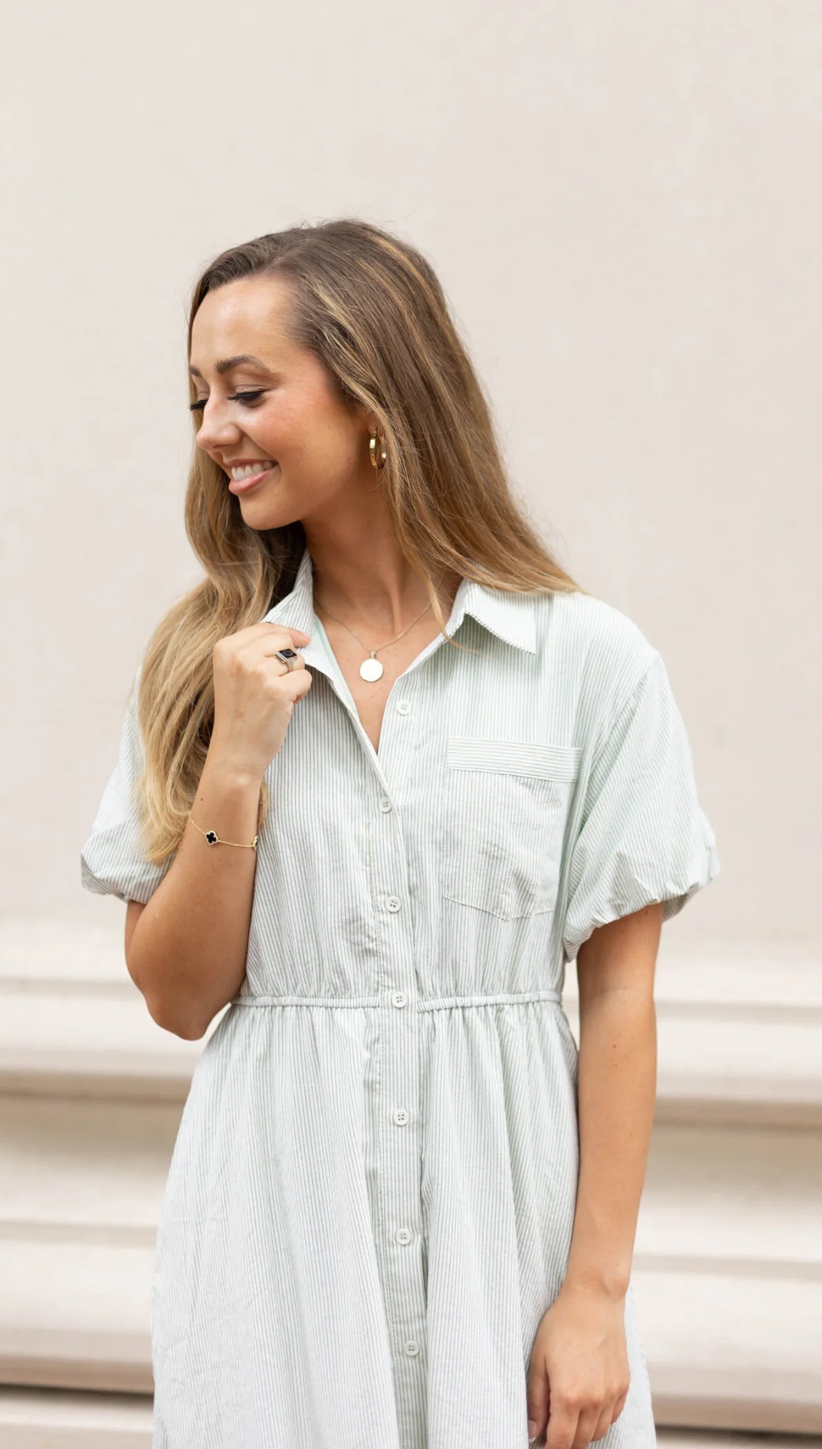 A person in a light green Carson Pin-Stripe Midi Dress by Dogwood Cloth, featuring buttons and an elastic waistband for added comfort and style, smiles while looking down. They are accessorizing with hoop earrings and a necklace.