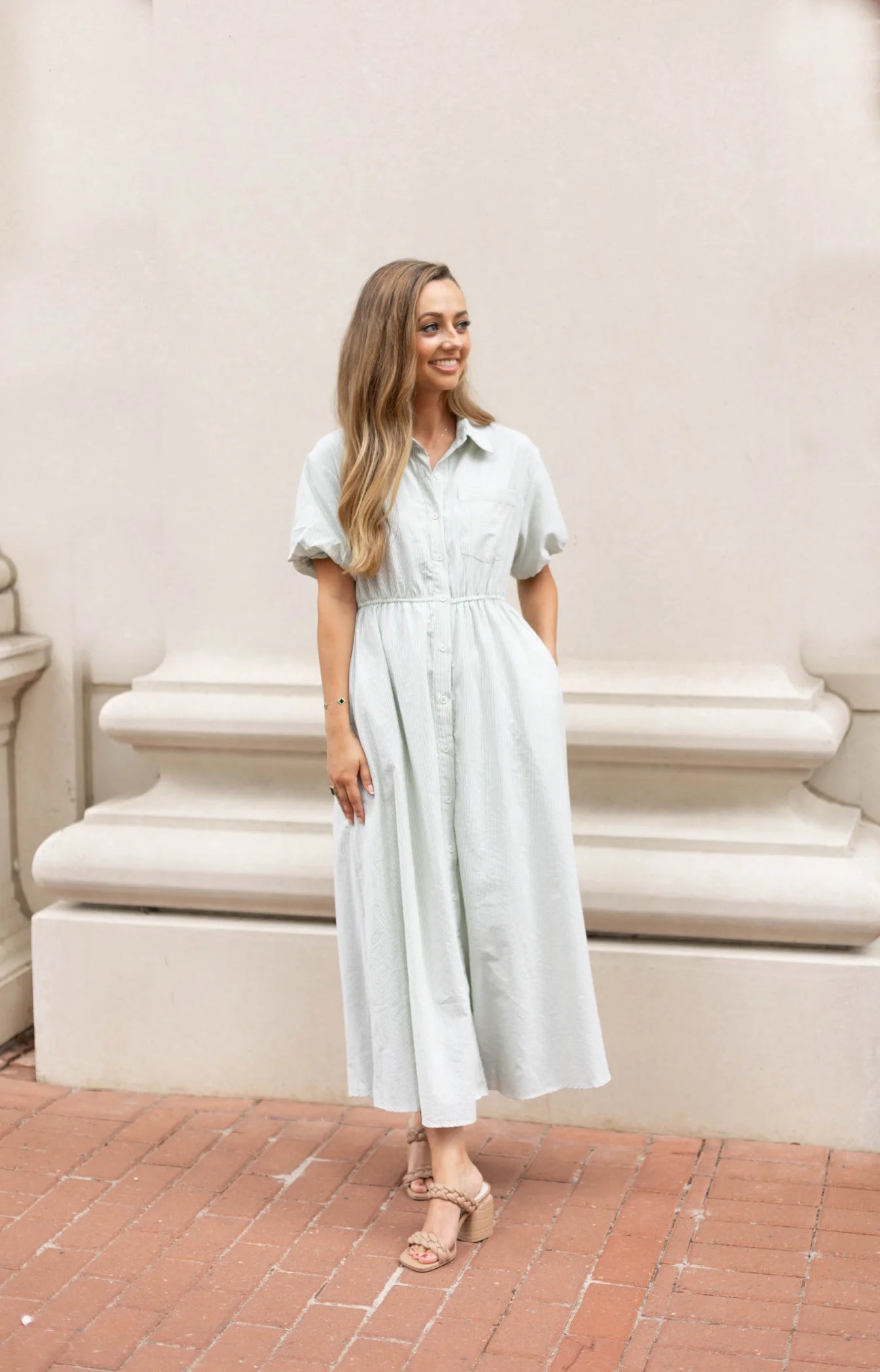 A woman wearing a Dogwood Cloth Carson Pin-Stripe Midi Dress in light green, featuring delicate pinstripes and an elastic waistband, stands on a brick path in front of a cream-colored wall.