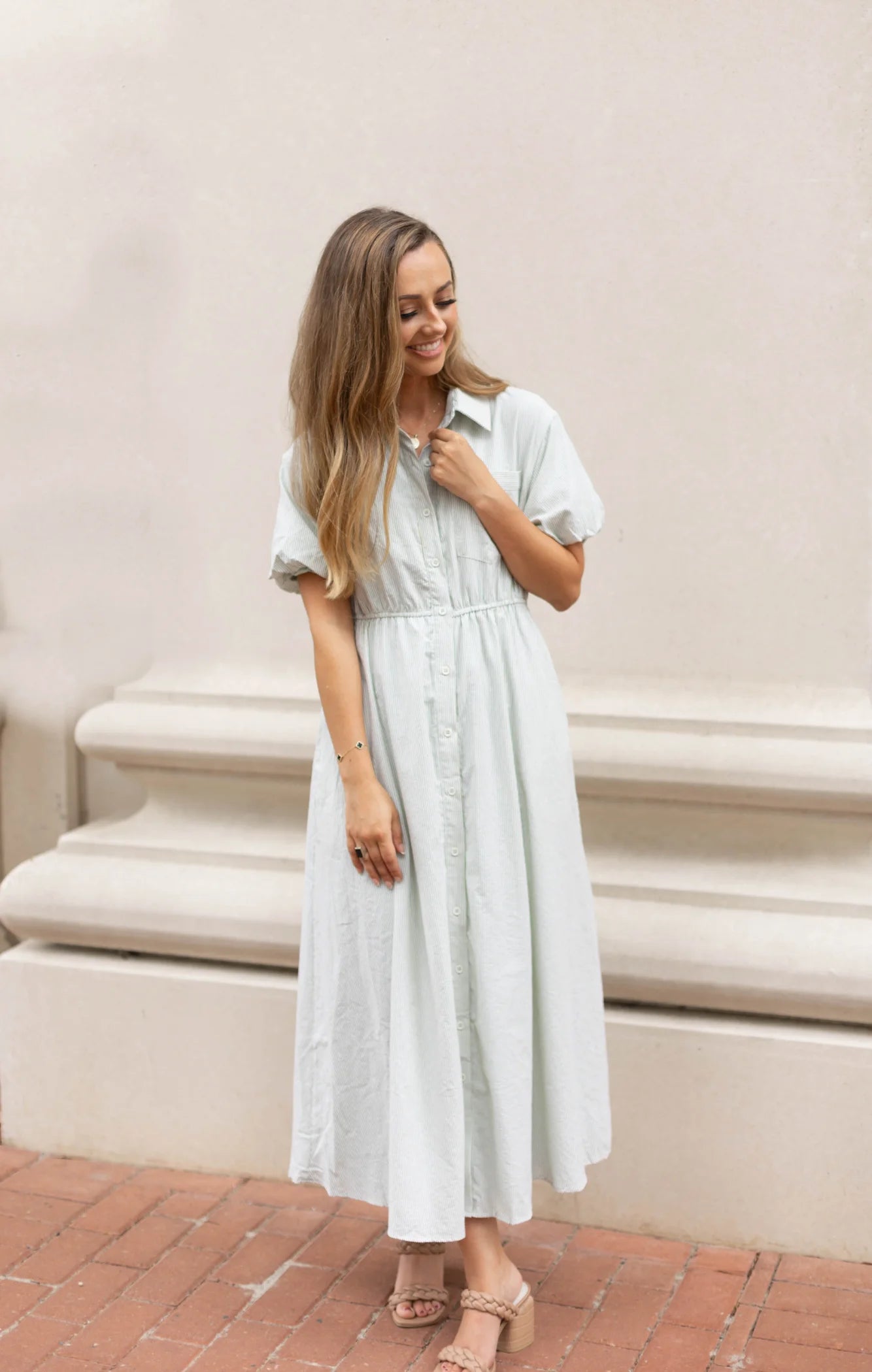 A person in a Carson Pin-Stripe Midi Dress by Dogwood Cloth stands smiling with hands together, in front of a white architectural structure on a brick walkway.