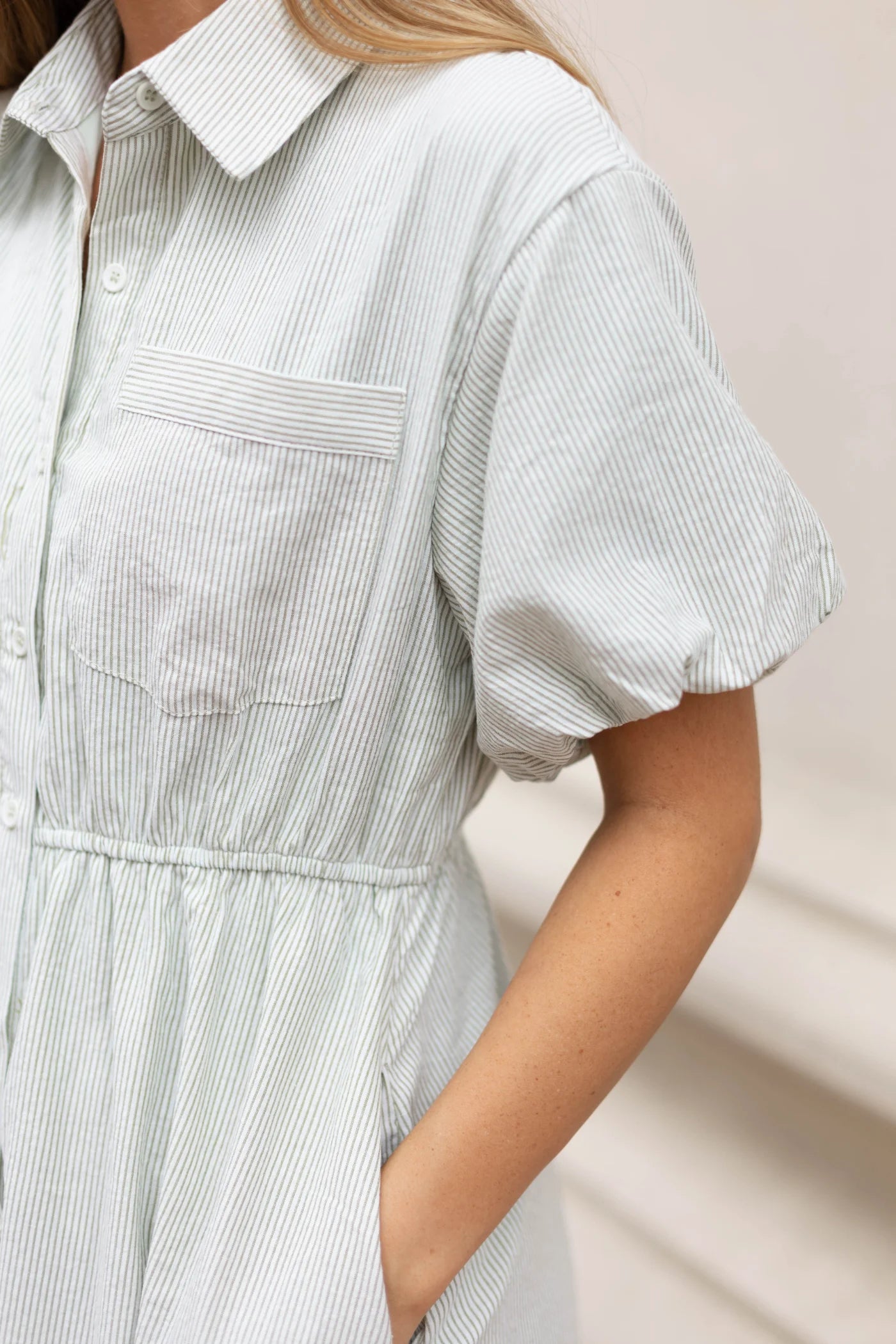 A close-up shows a person wearing the Dogwood Cloth Carson Pin-Stripe Midi Dress in light green, highlighted with subtle pinstripes. This short-sleeved piece includes an elastic waistband, a chest pocket, and handy side pockets.