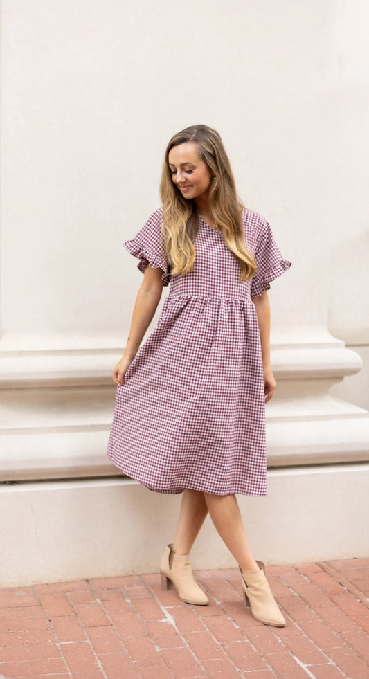 Clad in the Audrey Gingham Babydoll dress from Dogwood Cloth, a person wearing ankle boots poses elegantly against a light-colored wall, showcasing the burgundy gingham design with ruffle sleeves.