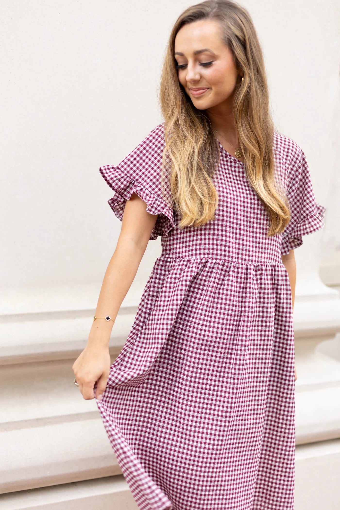 A person with long hair in an Audrey Gingham Babydoll dress from Dogwood Cloth stands against a white background, gracefully holding the dress with one hand.