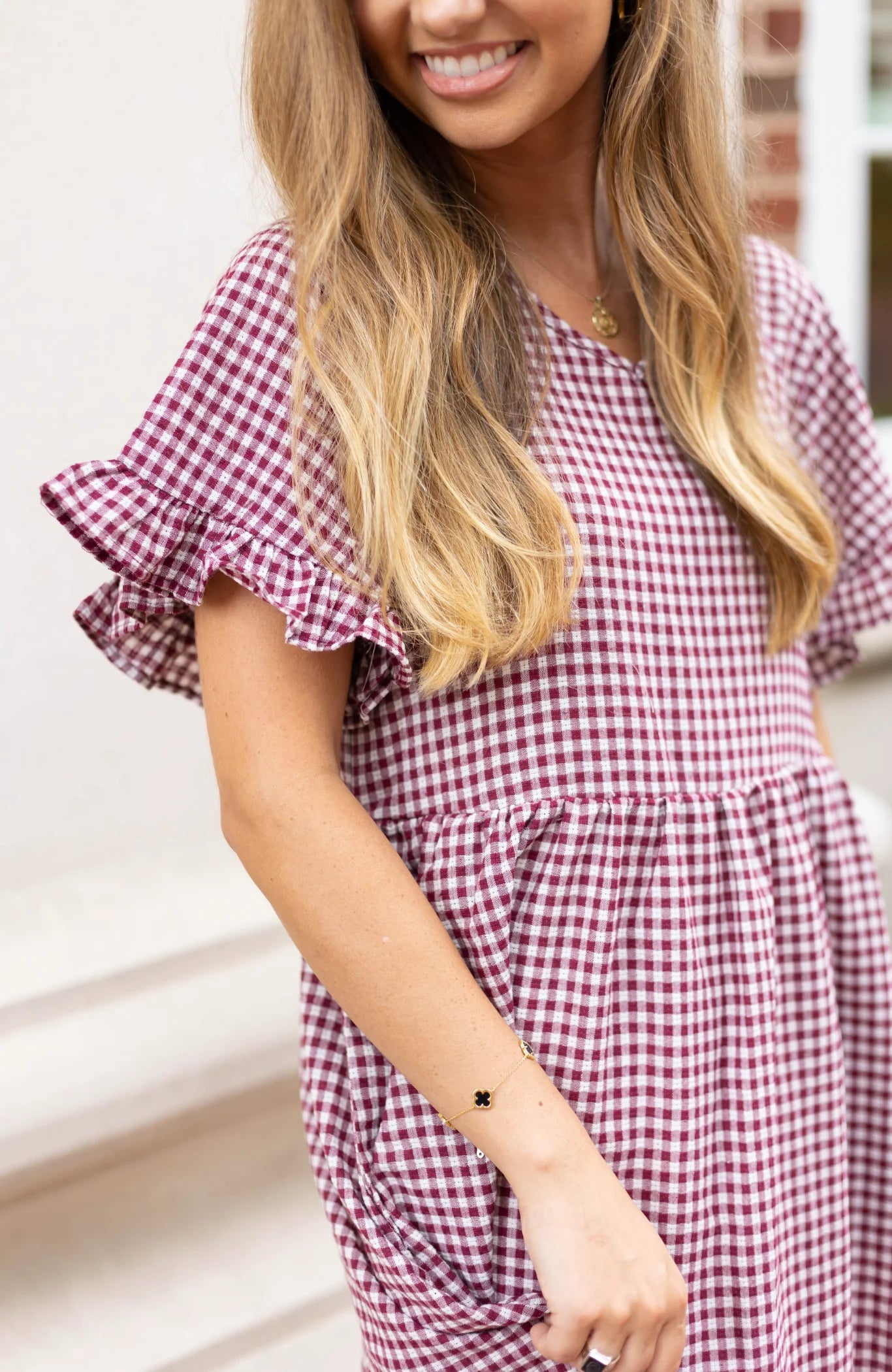 A woman standing outdoors and smiling is wearing the Audrey Gingham Babydoll dress by Dogwood Cloth, featuring burgundy gingham fabric and ruffle sleeves.