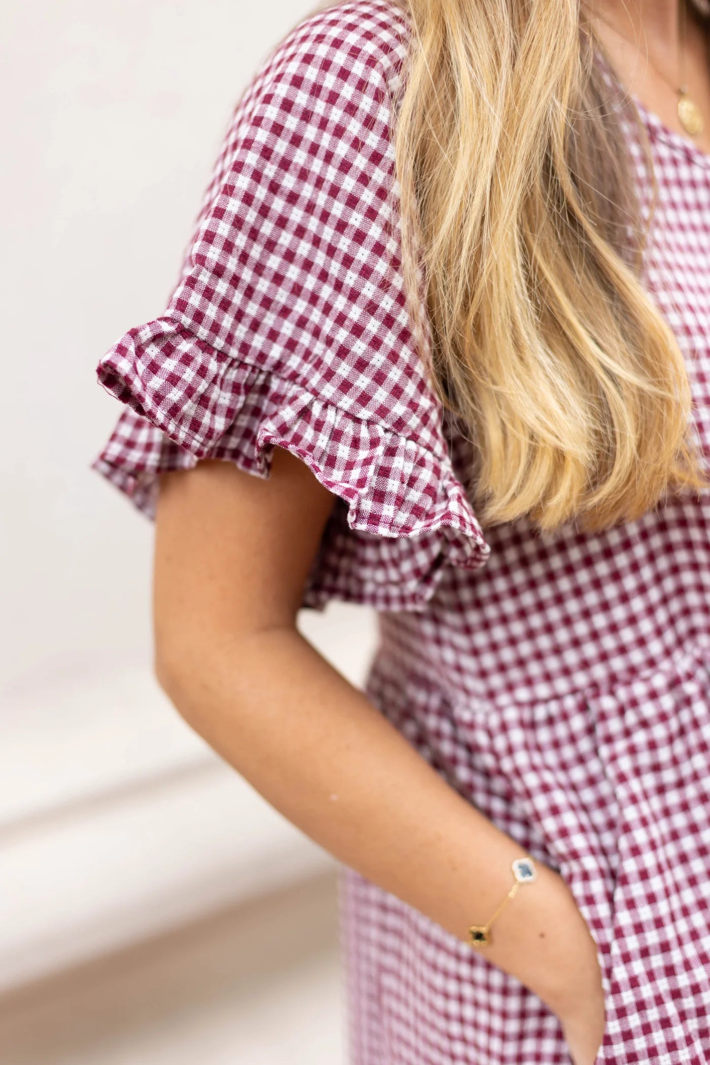 A close-up of a person wearing the Audrey Gingham Babydoll by Dogwood Cloth in burgundy showcases its ruffle sleeves. Their long blonde hair cascades down, complemented by a bracelet on their wrist, adding a feminine touch to the ensemble.