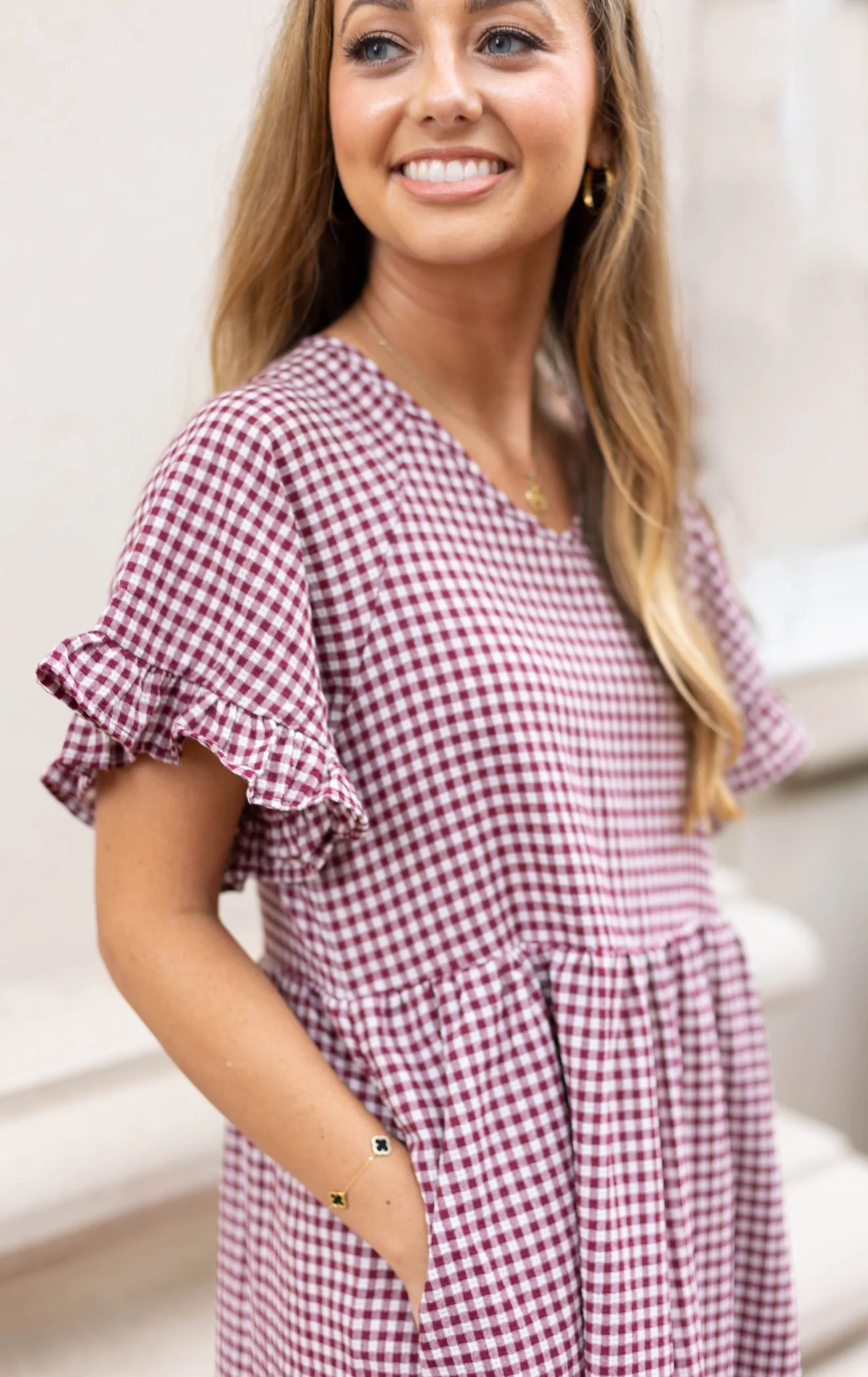 A woman smiles warmly, showcasing the Audrey Gingham Babydoll by Dogwood Cloth, a dress with a feminine touch in burgundy. The ruffle sleeves add charm as she stands against a light background.