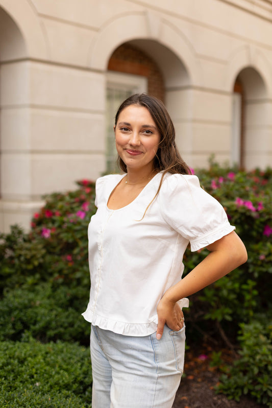 A woman with long brown hair stands outdoors in front of bushes and a building with arched windows, wearing the Cory Top by Dogwood Cloth and light blue jeans.