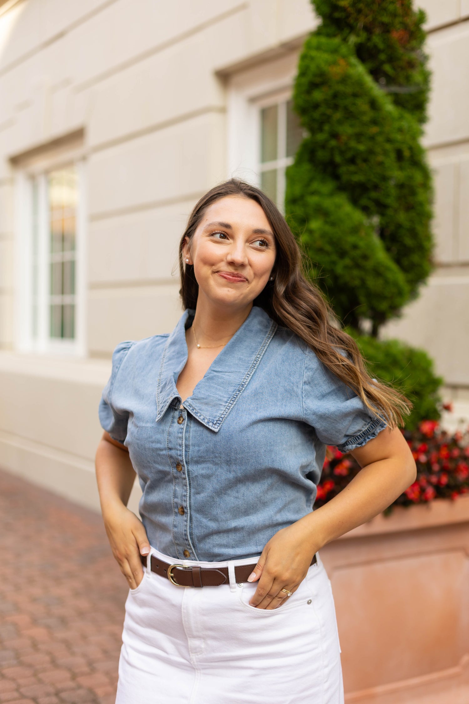 Wearing the Dogwood Cloth Jessa Jean Top and a white skirt, a woman stands outdoors before a building and greenery, smiling slightly and glancing to the side.