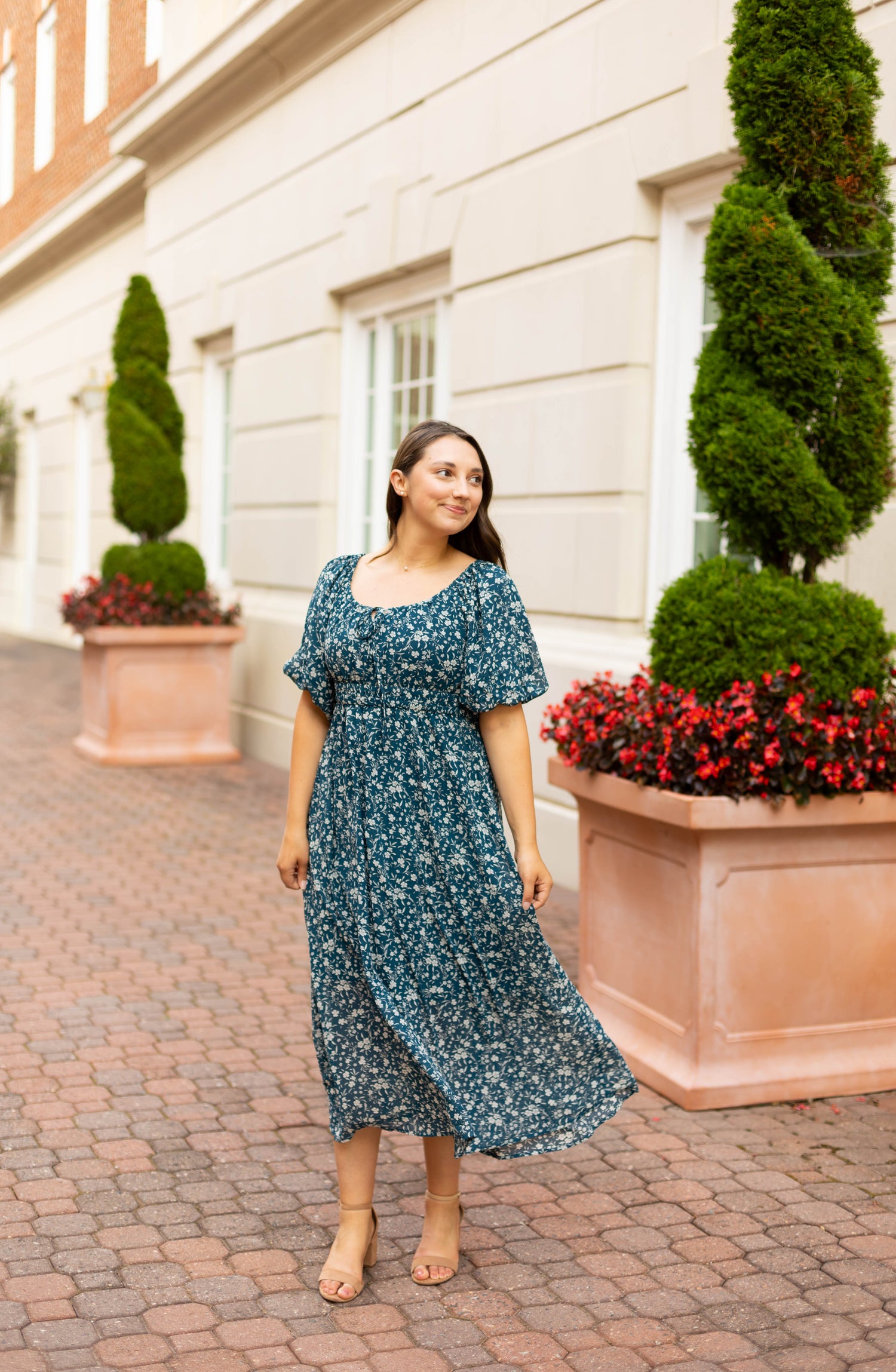 A woman in the Dogwood Cloth Aubrey Midi Dress, featuring a blue floral print, stands on a brick walkway near large potted plants and a cream-colored building.