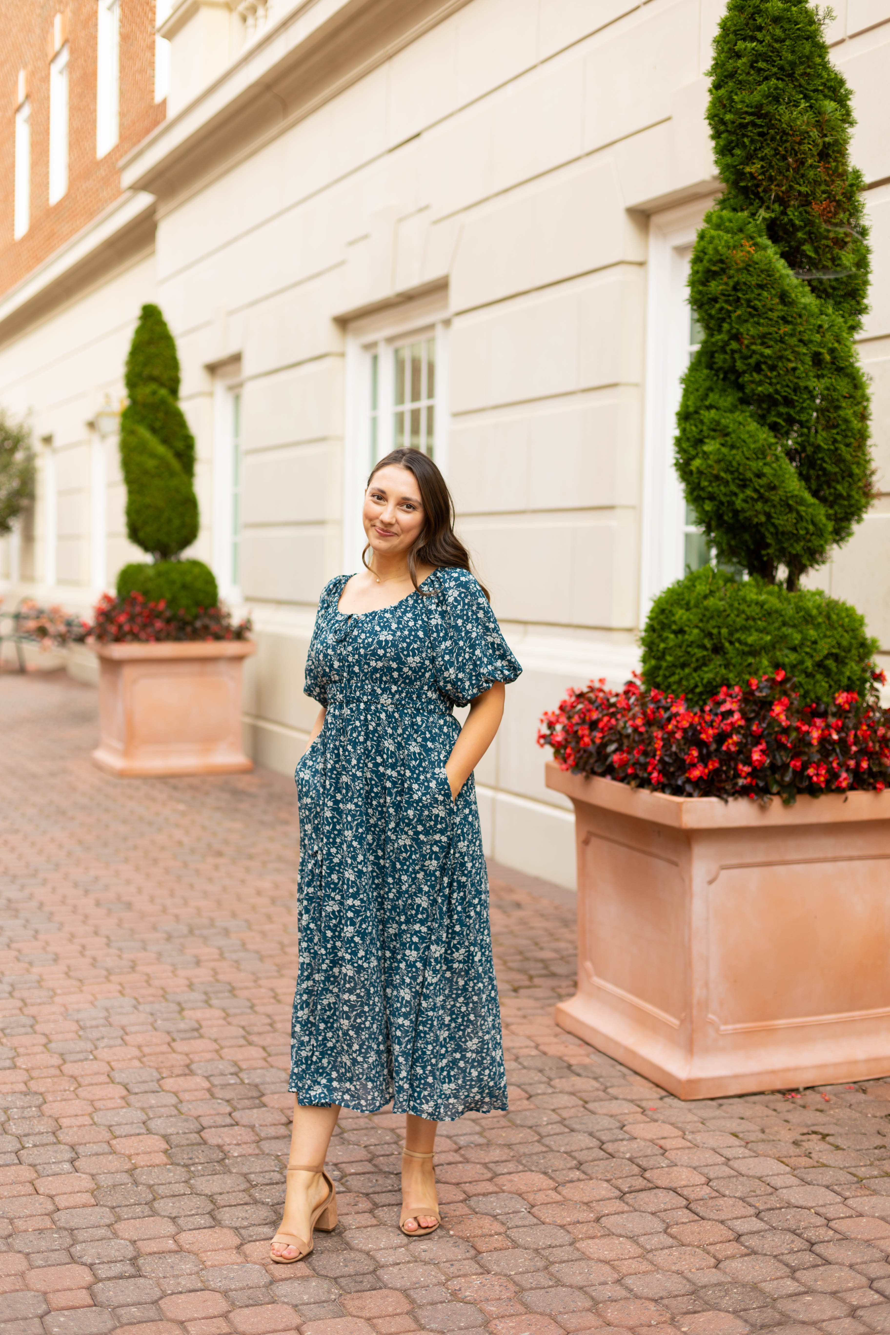 A woman in the elegant Dogwood Cloth Aubrey Midi Dress with an empire waist stands on a brick walkway between planters with shaped trees and red flowers, in front of a beige building.