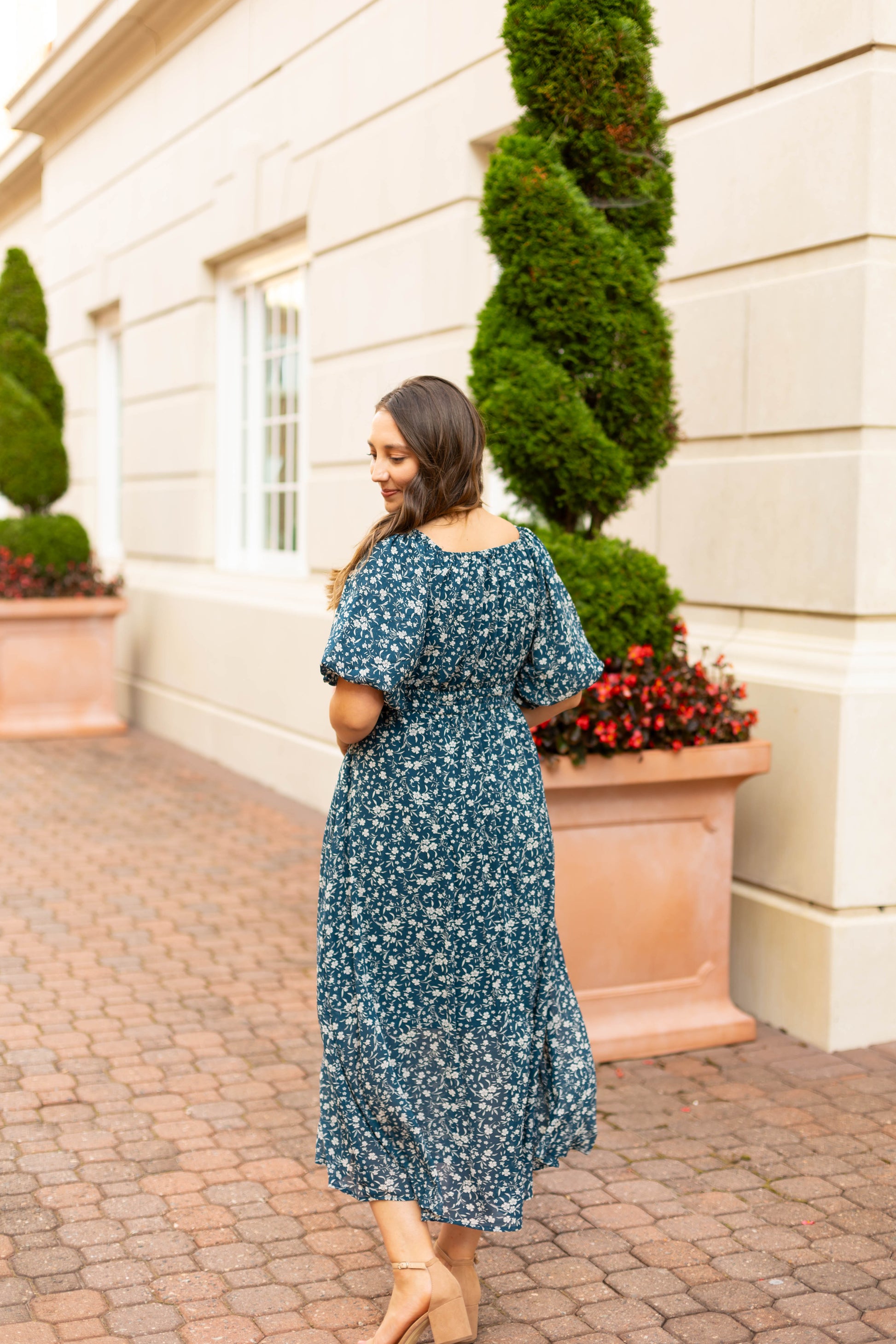 A woman in the elegant blue floral Aubrey Midi Dress by Dogwood Cloth, featuring an empire waist, walks along a brick path beside a light building with large planters and manicured shrubs.