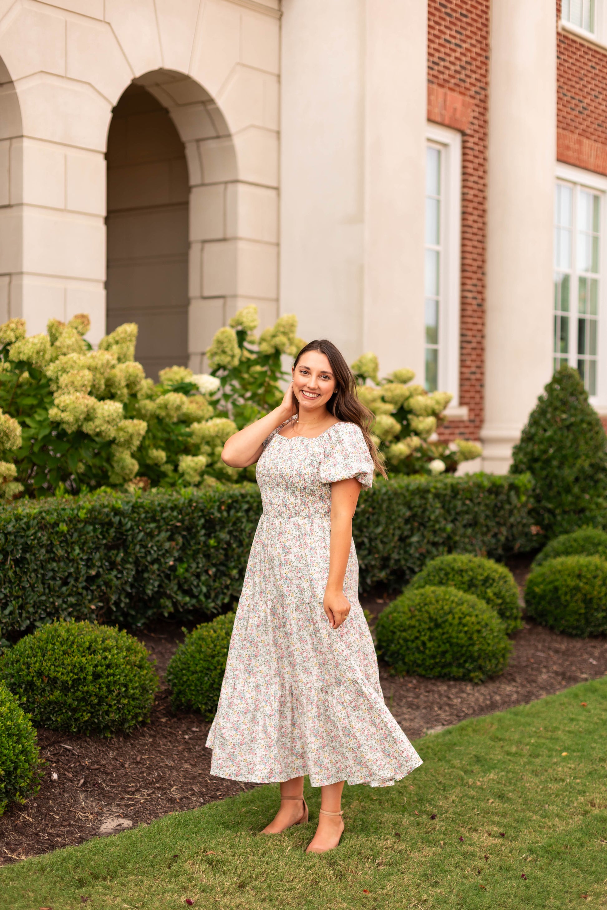 Wearing the Meadow Tiered Maxi by Dogwood Cloth, a woman poses and smiles on a lawn near bushes and hydrangeas in front of a brick and stone building.