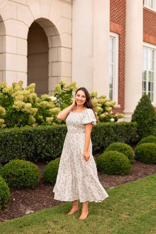 Wearing the Meadow Tiered Maxi by Dogwood Cloth, a woman poses and smiles on a lawn near bushes and hydrangeas in front of a brick and stone building.