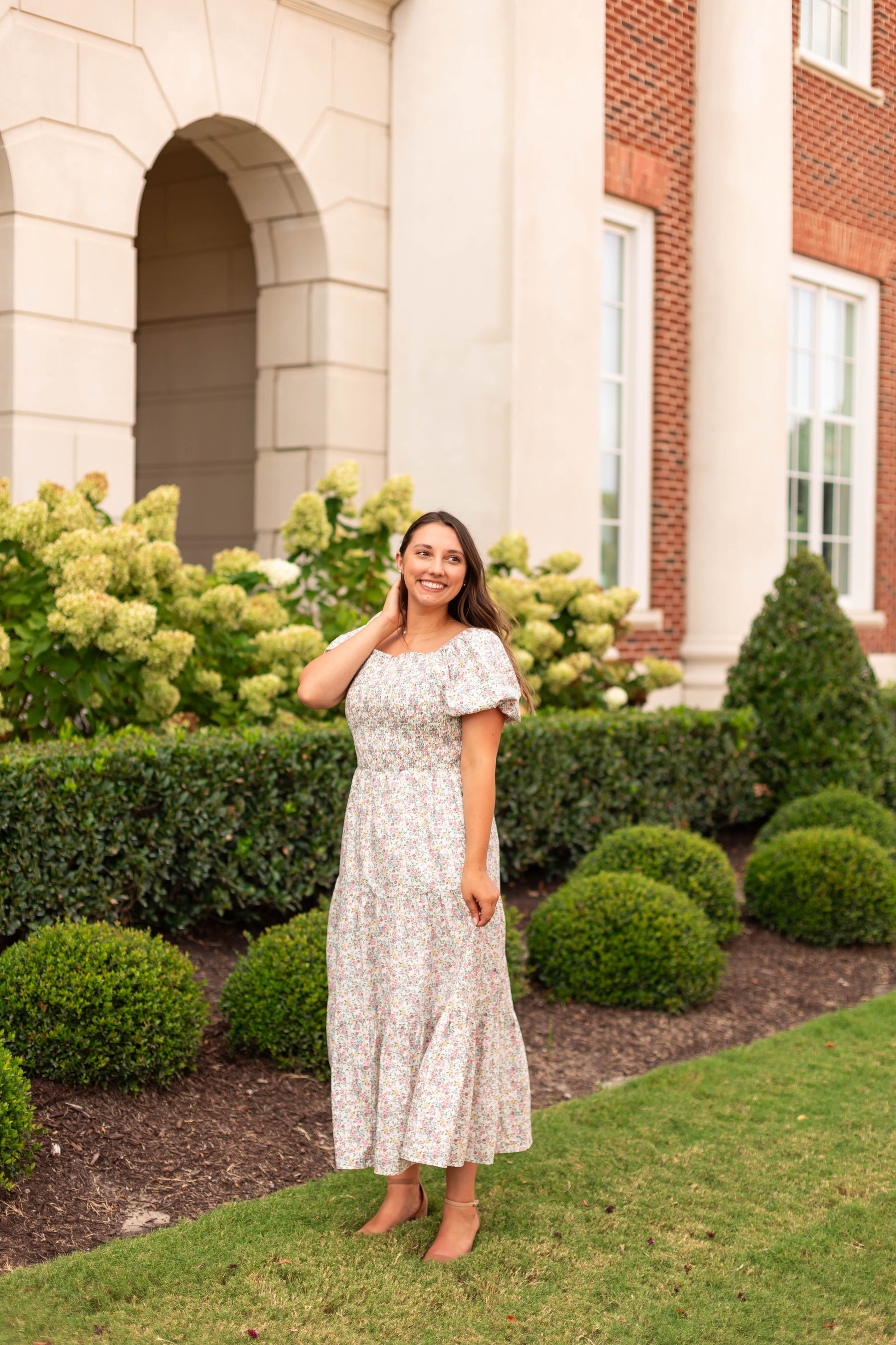 A woman wears the Meadow Tiered Maxi by Dogwood Cloth, standing on a lawn with large windows, white columns, and landscaped bushes in the background.