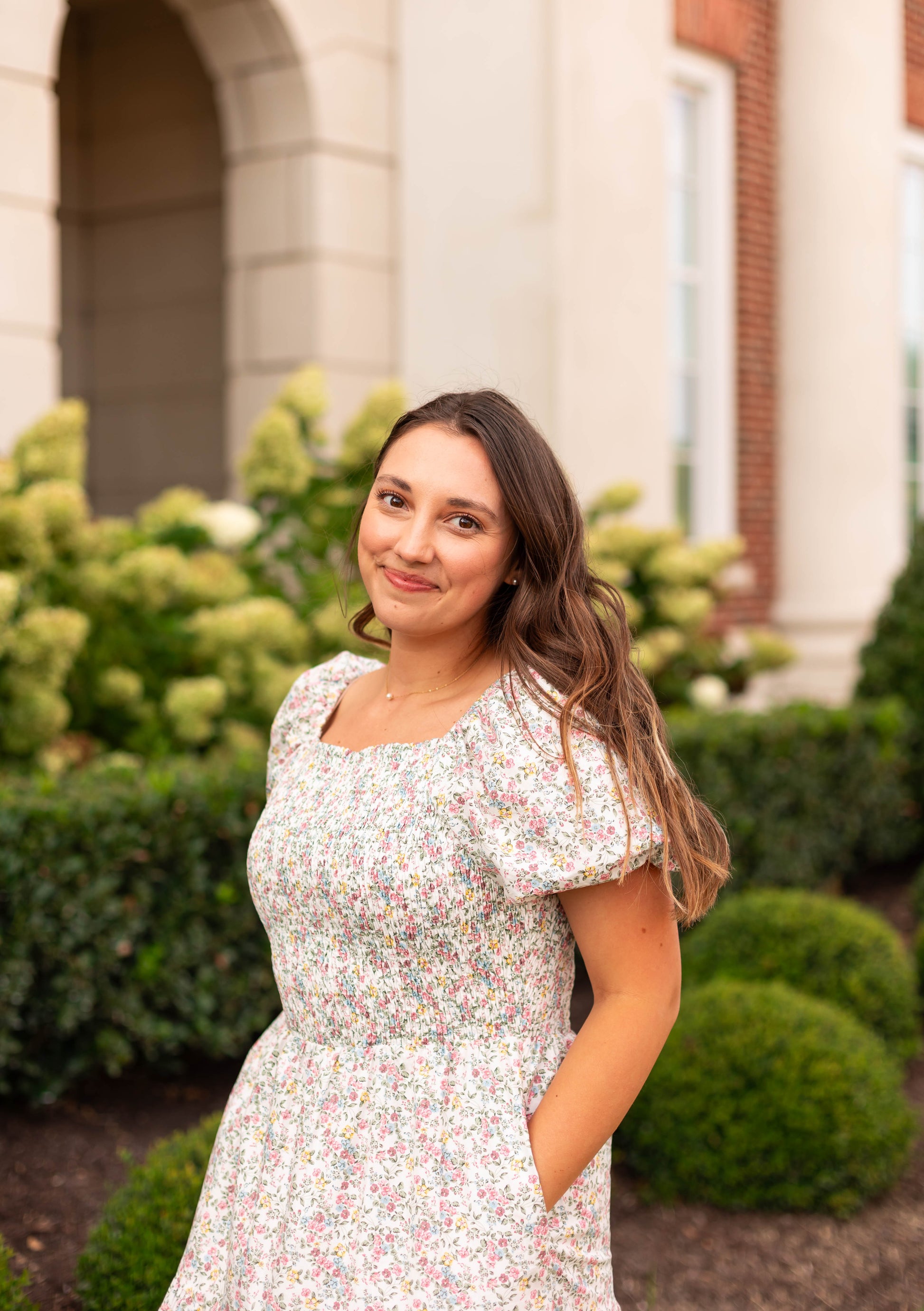 A woman wears the Meadow Tiered Maxi by Dogwood Cloth, standing outdoors before a building with lush greenery and bushes in the background.