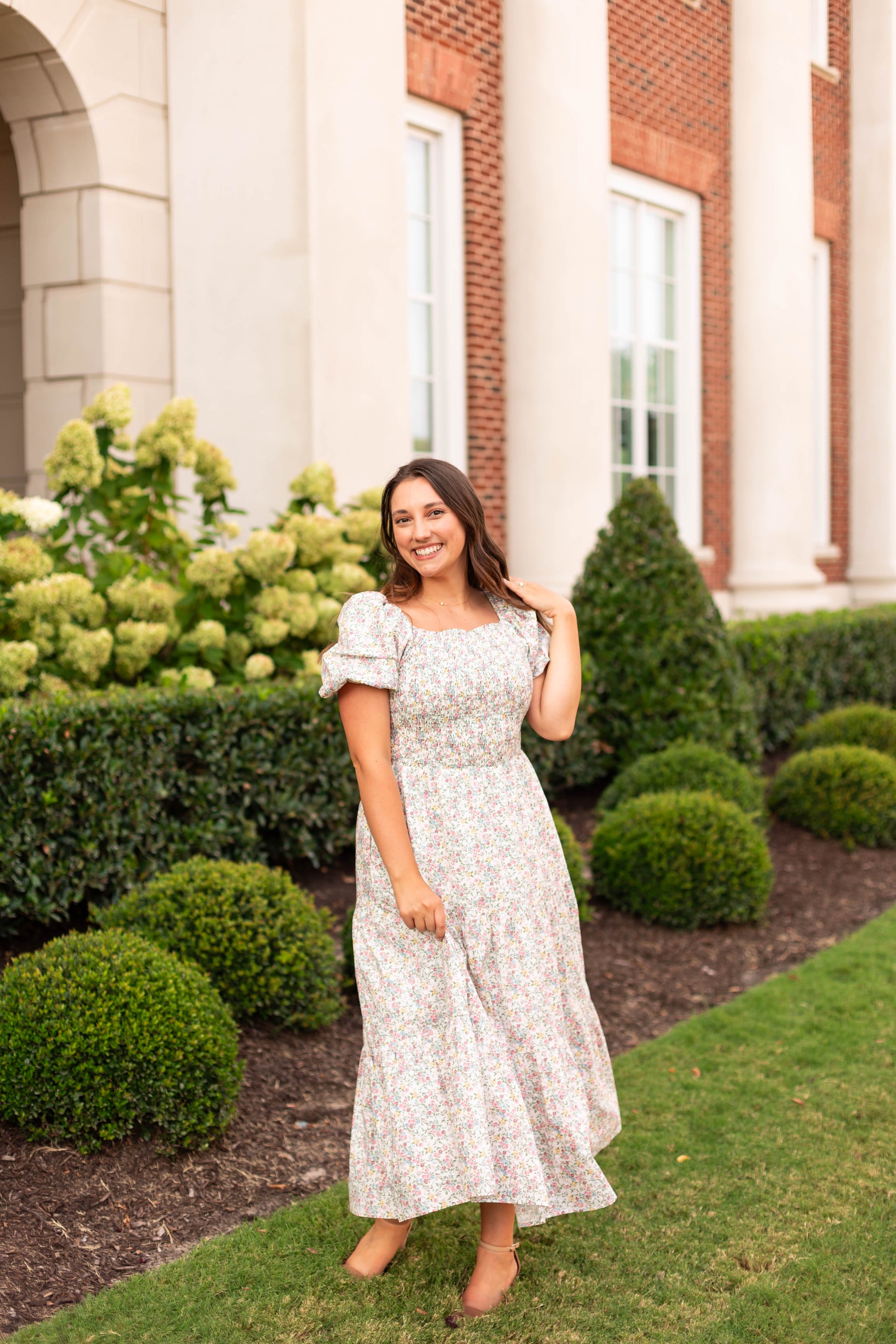 A woman in the Dogwood Cloth Meadow Tiered Maxi smiles on a lawn in front of a brick building with large windows and manicured bushes.