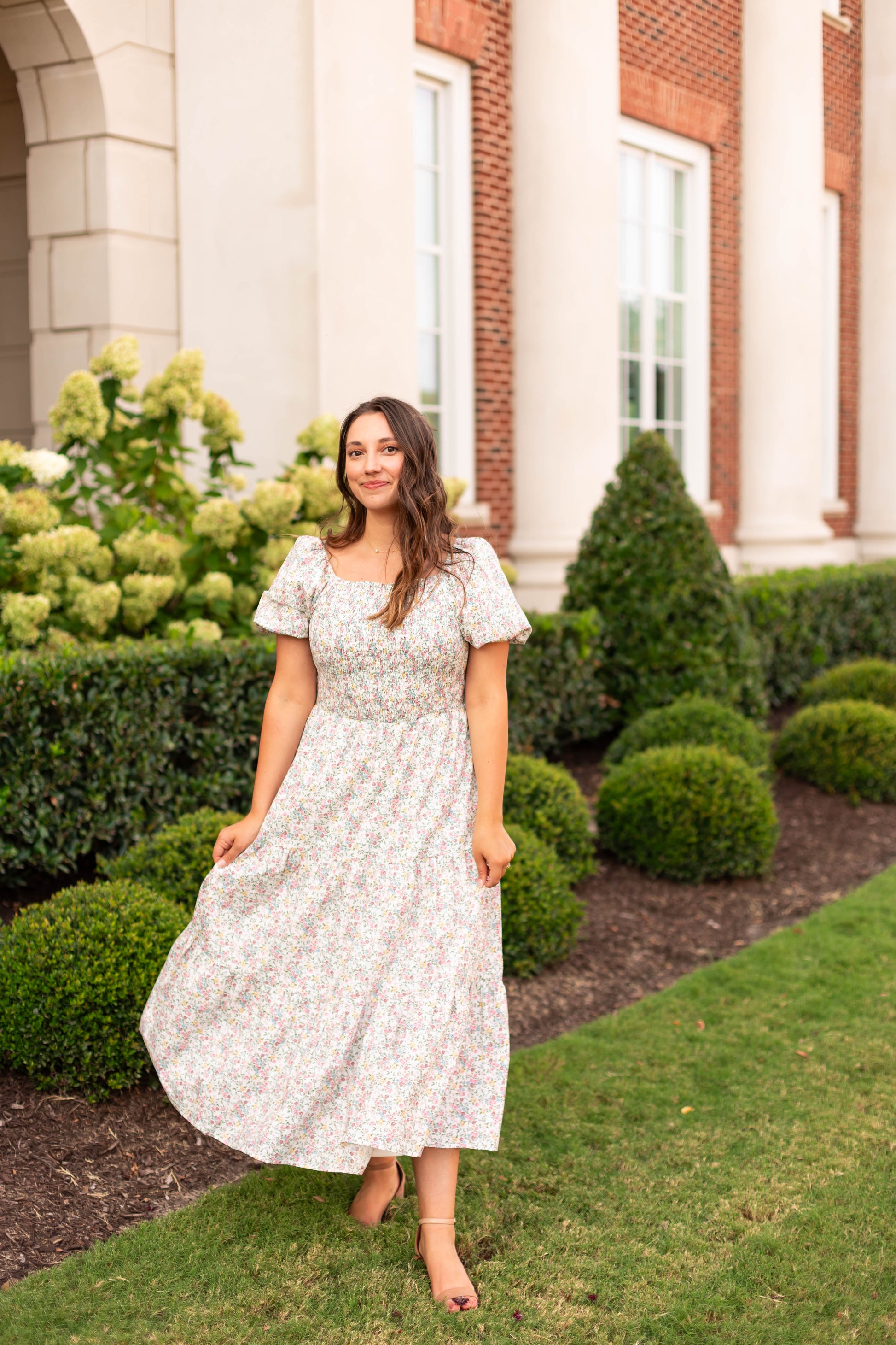 A woman wearing the Dogwood Cloth Meadow Tiered Maxi stands on a lawn before a columned building and manicured bushes.
