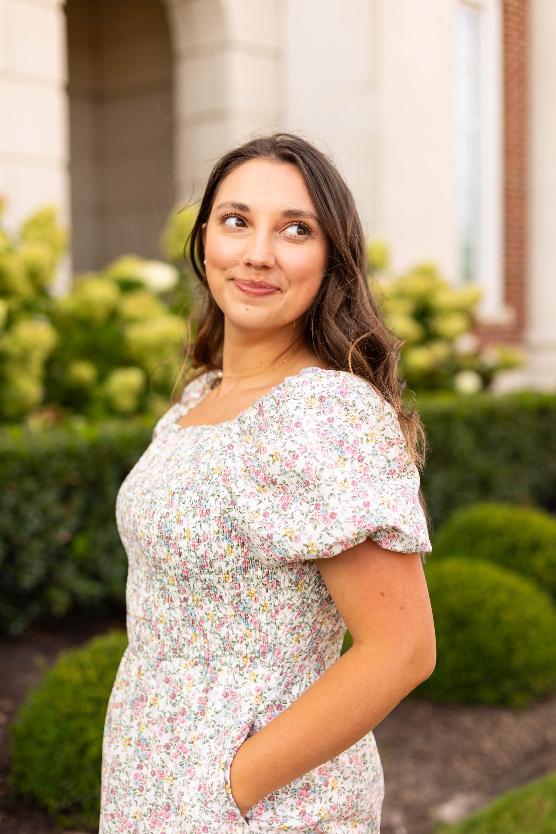 A woman wearing the Dogwood Cloth Meadow Tiered Maxi stands outdoors with one hand in her pocket, looking to the side. Green shrubs and a building appear in the background.
