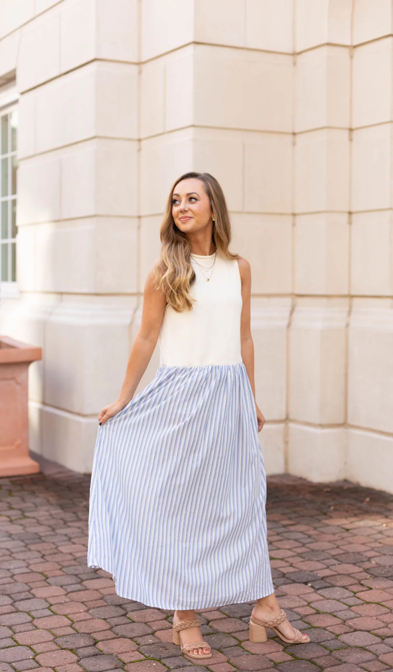 A woman in the Mia Contrast Midi by Dogwood Cloth stands elegantly on a cobblestone path, near a beige building.