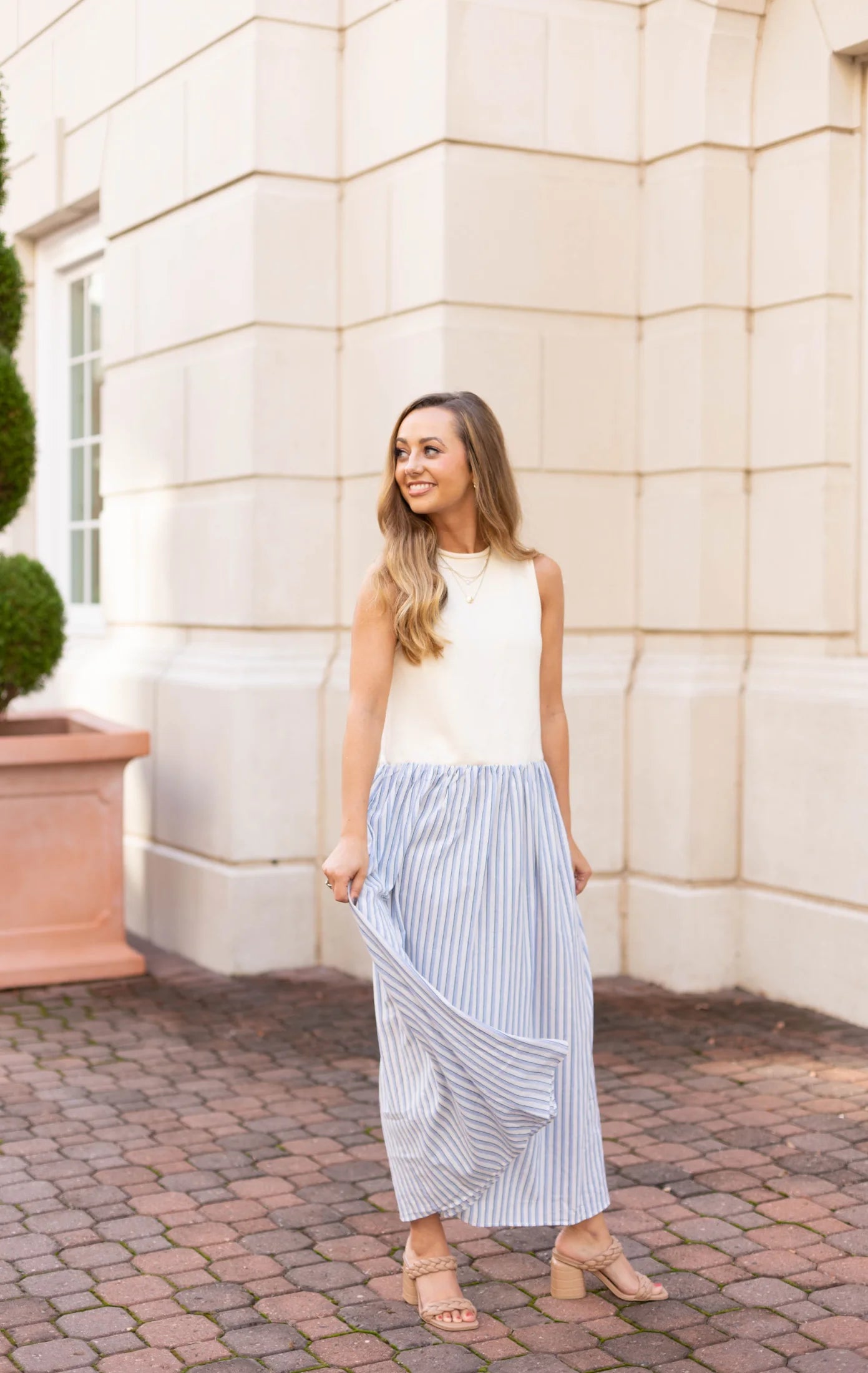 A woman wearing the Mia Contrast Midi by Dogwood Cloth, featuring a sleeveless sweater bodice and a striped skirt, stands on a cobblestone path, smiling with a beige building in the background.