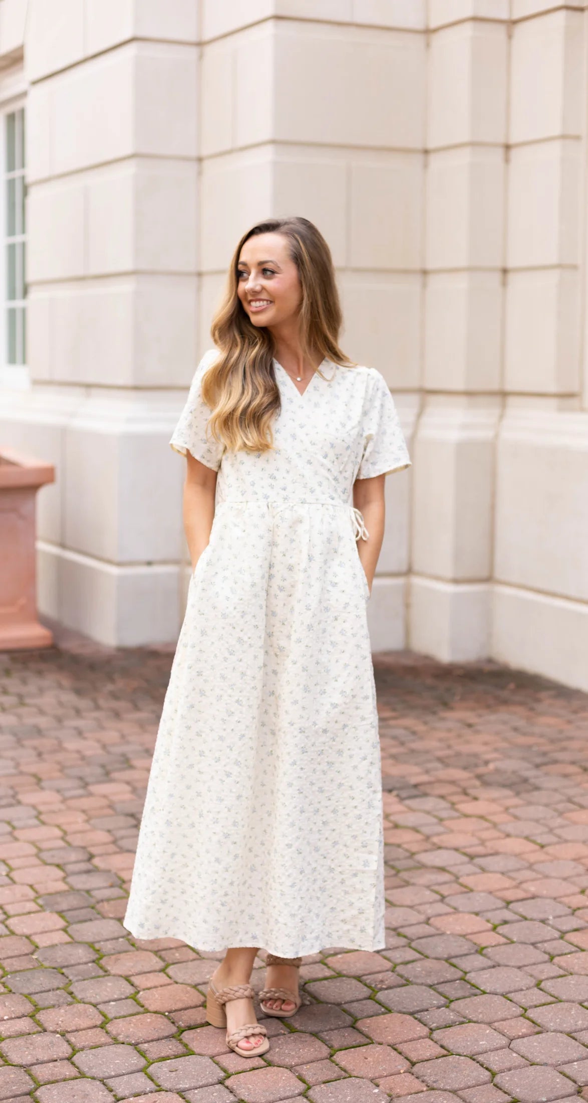 A woman in a Dogwood Cloth Grace Wrap Dress stands on a cobblestone path in front of a stone building, with its elegant floral pattern accentuating its maternity-friendly design.