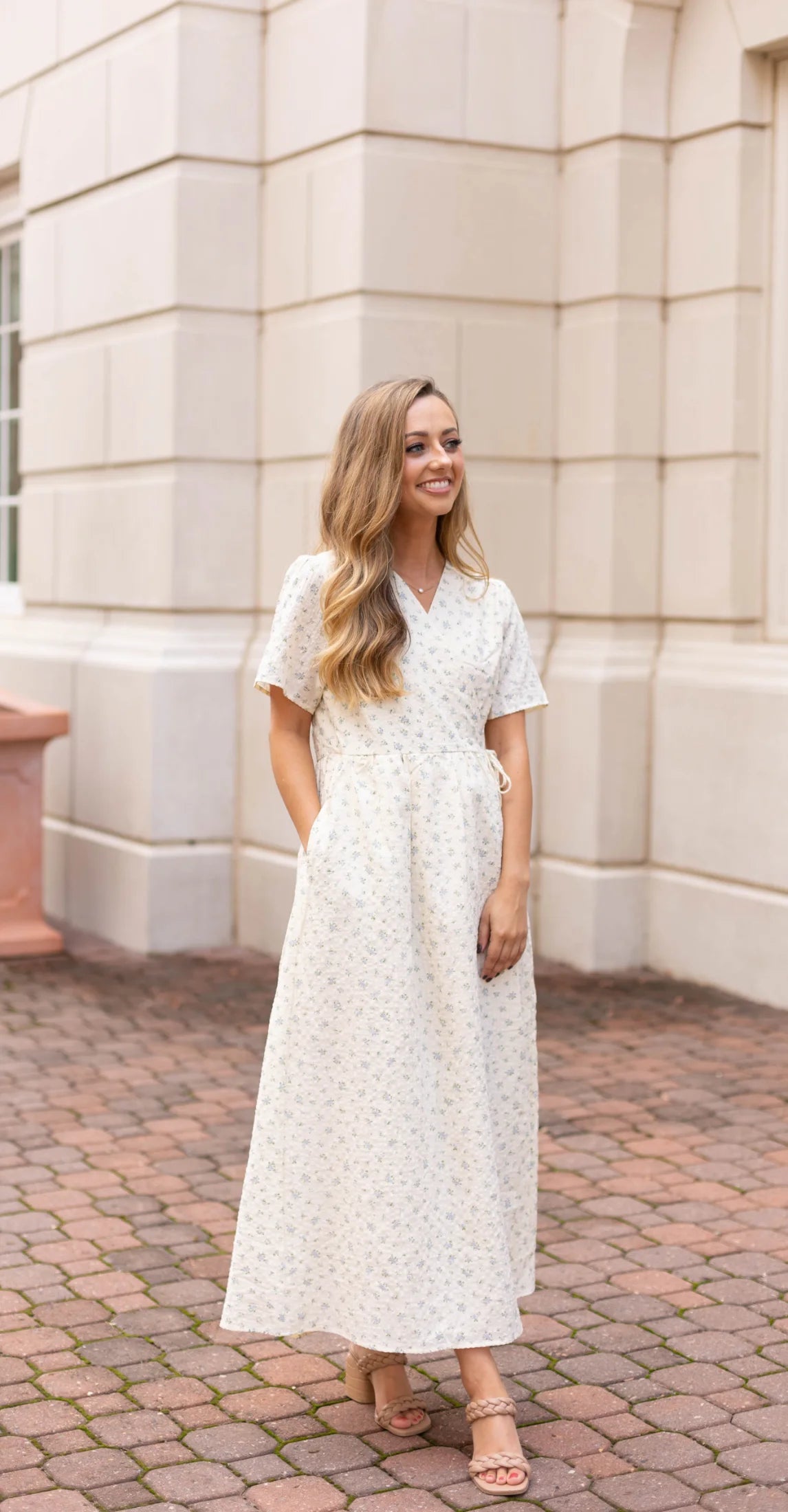 A woman in a Dogwood Cloth Grace Wrap Dress, showcasing a delightful floral design, stands on a brick path in front of a beige building.