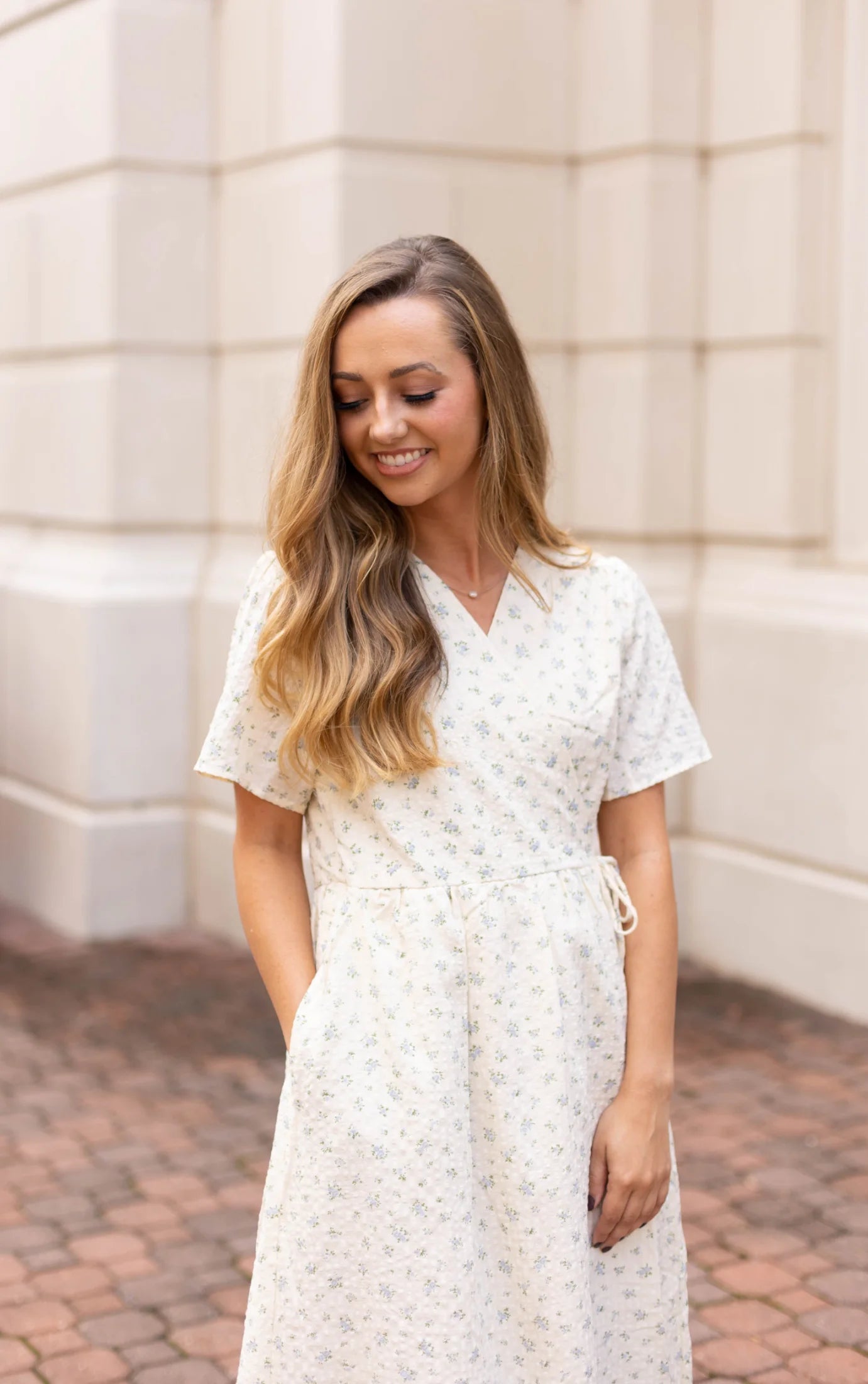 A woman with long hair stands on a brick walkway, wearing the Grace Wrap Dress by Dogwood Cloth. This short-sleeve, light-colored piece features a vibrant floral pattern, making it ideal for maternity-friendly styling. She smiles as she places her hands in the dress's pockets.