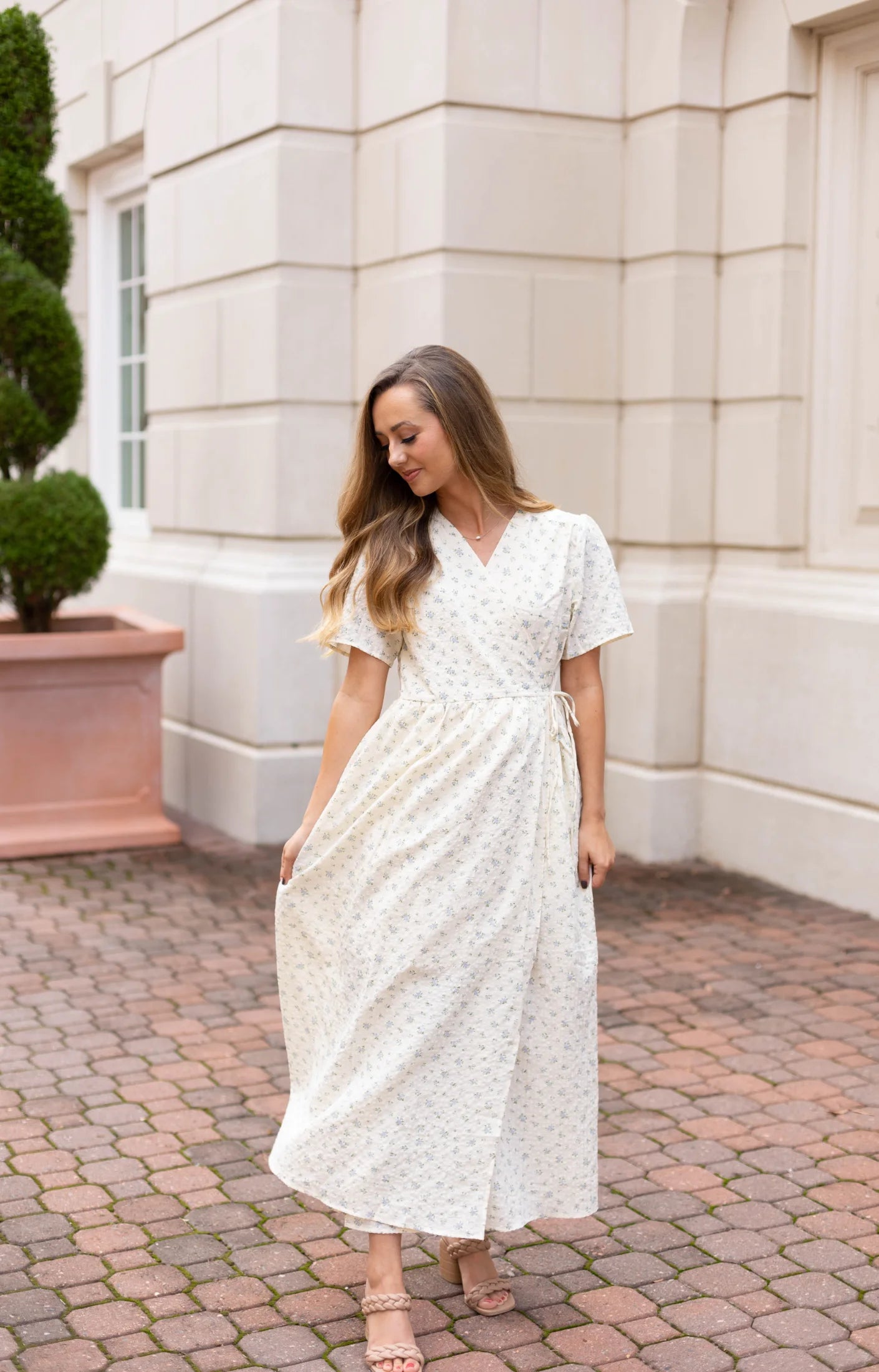 A woman wearing a Grace Wrap Dress by Dogwood Cloth, suitable for maternity wear and featuring an elegant floral design, poses on a brick path in front of a light-colored building with greenery-filled planters.