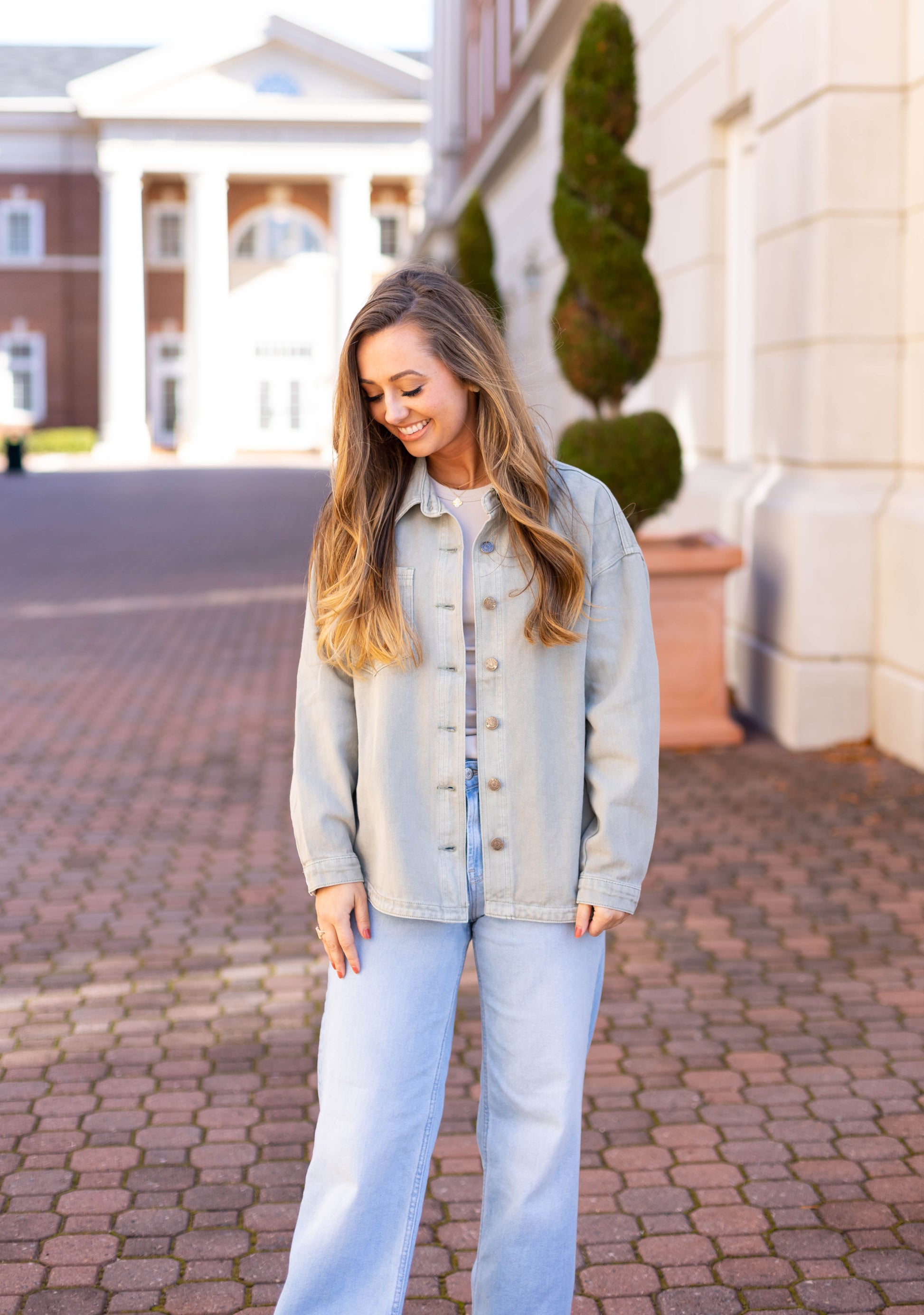 Wearing the Leah Denim Jacket by Dogwood Cloth, a woman stands smiling on a brick pathway. A building with columns in the background adds an elevated style to this ultimate wardrobe essential scene.