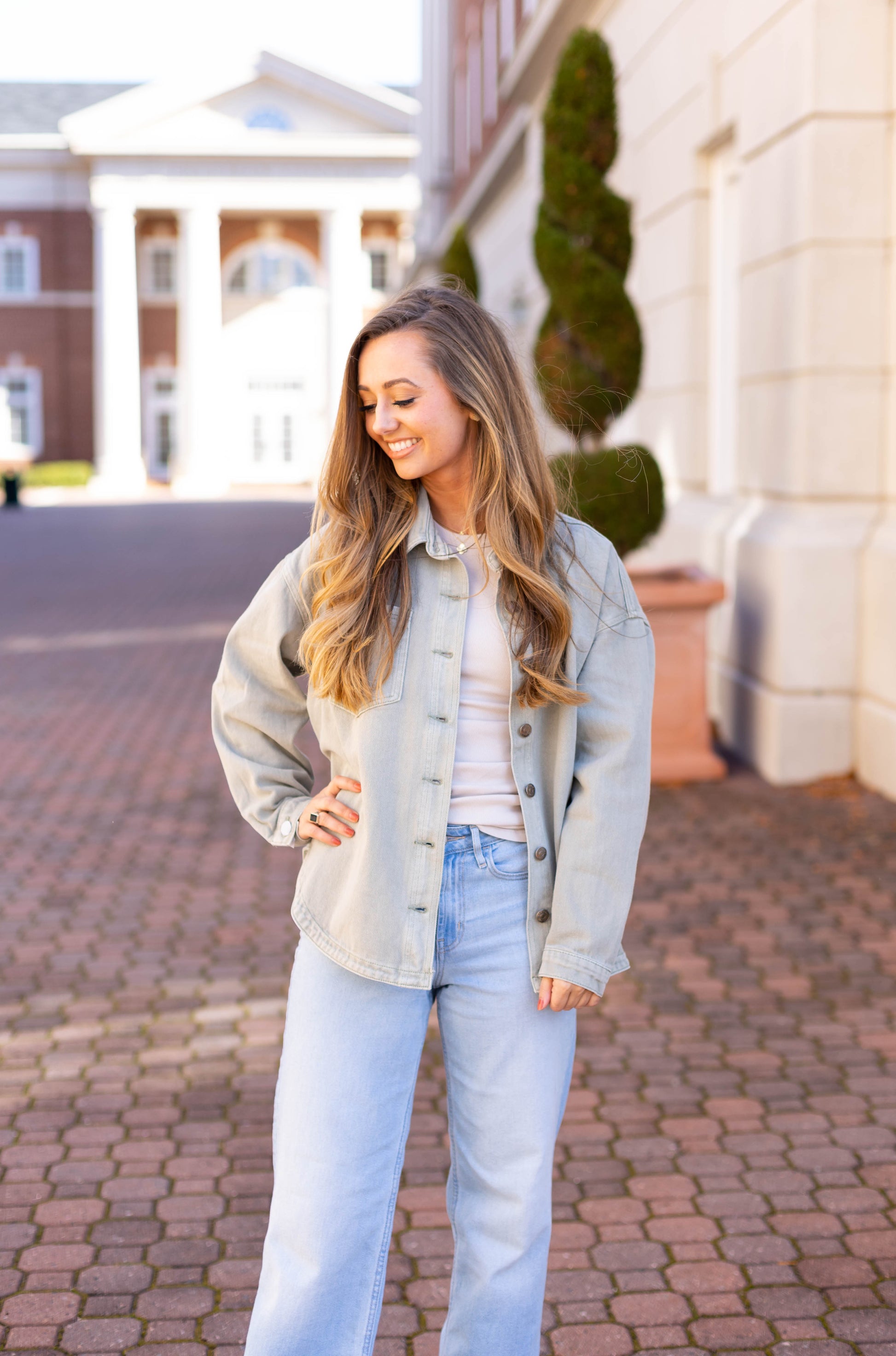 A person with long hair, wearing Dogwood Cloth's Leah Denim Jacket and jeans, stands on a brick pathway before a columned building, looking to the side and smiling.