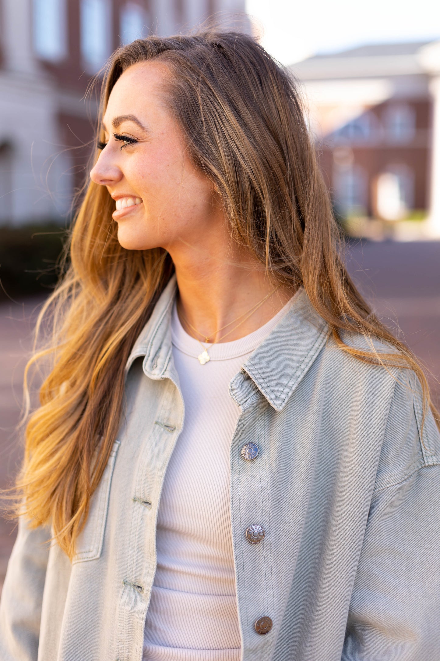 A woman with long hair smiles and looks to her left, showcasing a versatile style in a light denim jacket and the Dogwood Cloth Marie Everyday Top while standing outdoors.