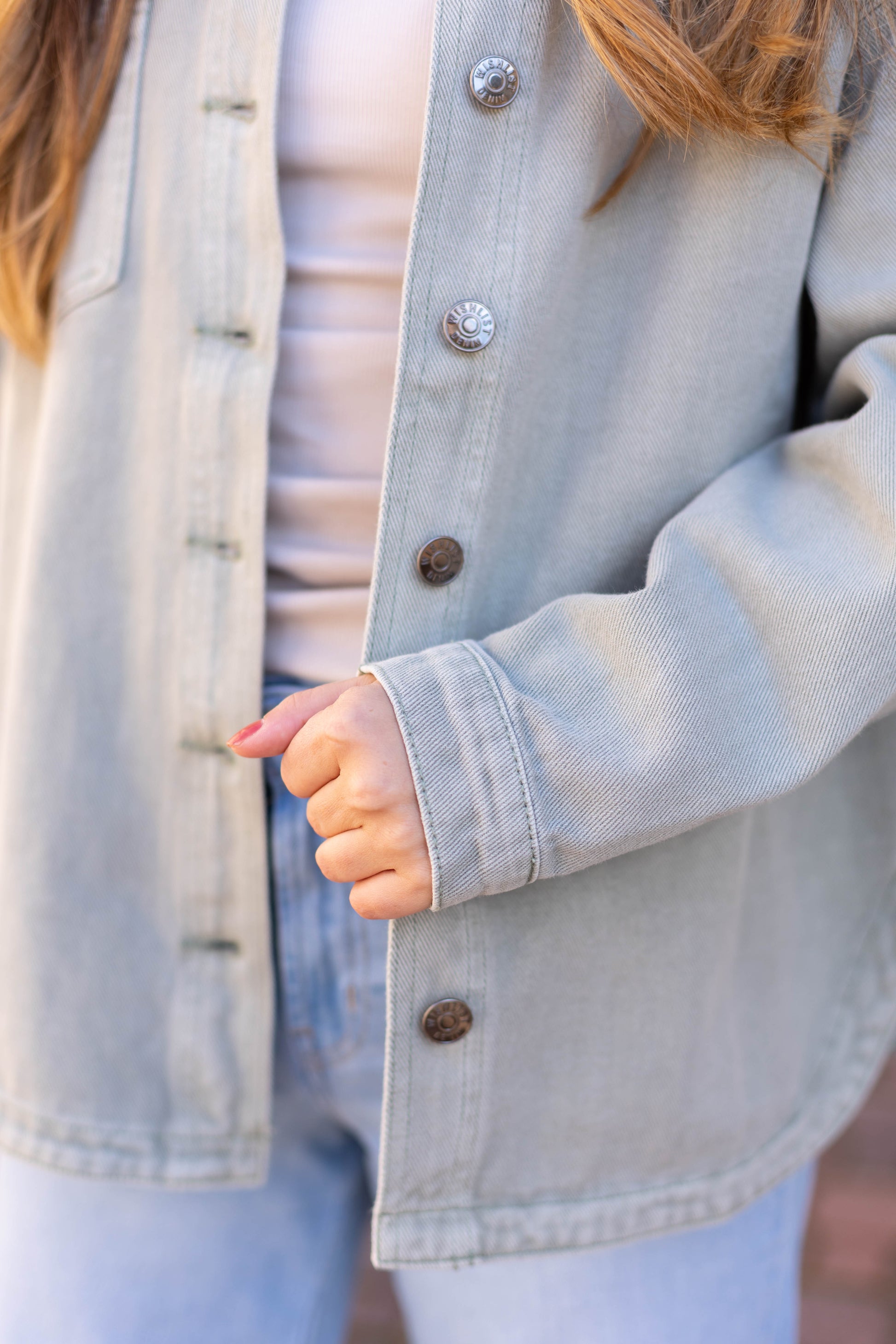 A person elevates their style in the Leah Denim Jacket by Dogwood Cloth, layering it over a white top and blue jeans while deftly pulling the jacket's edge with their left hand.