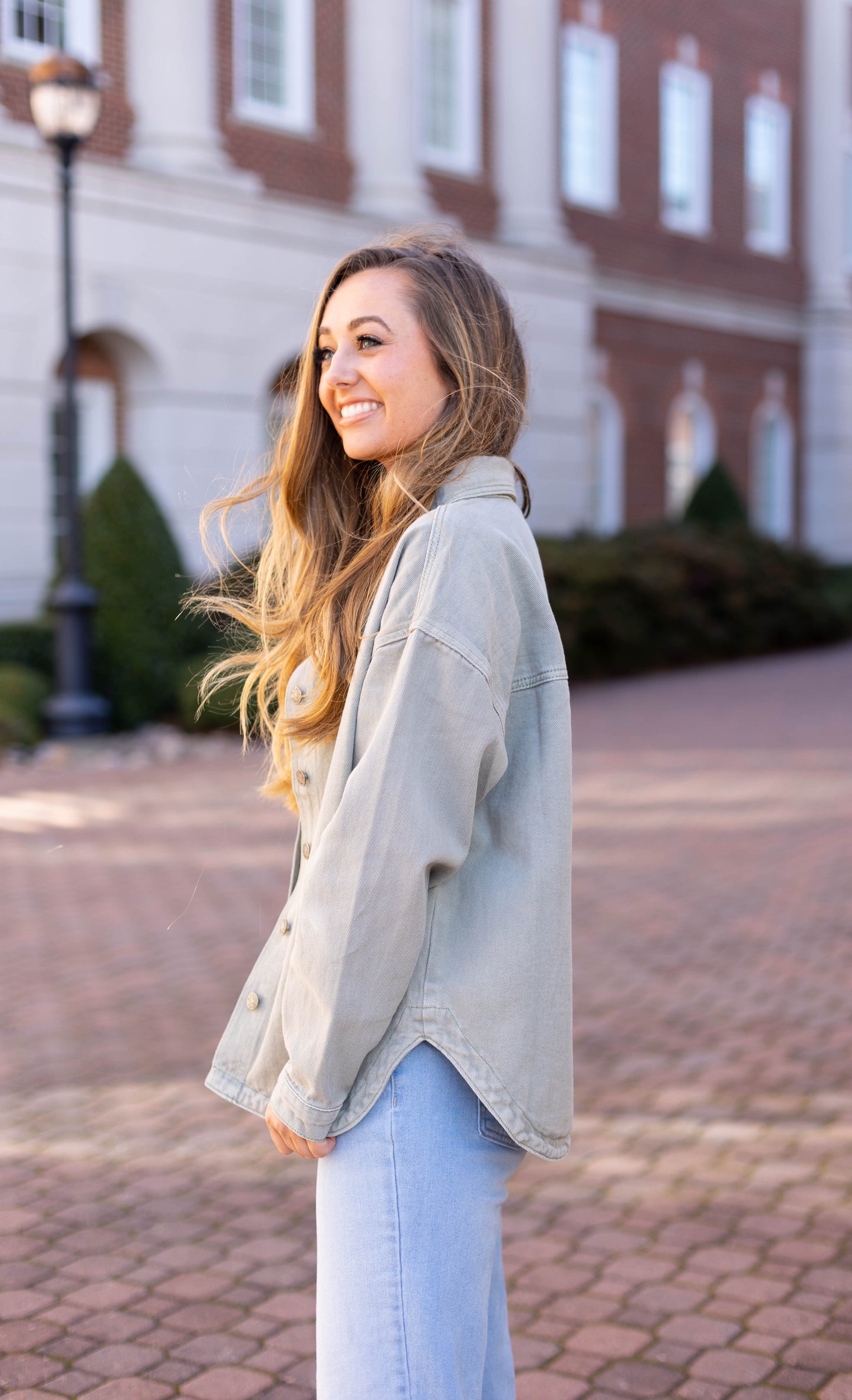 A woman wearing a Leah Denim Jacket by Dogwood Cloth smiles while standing on a brick pathway with a building in the background, showcasing this essential wardrobe piece.