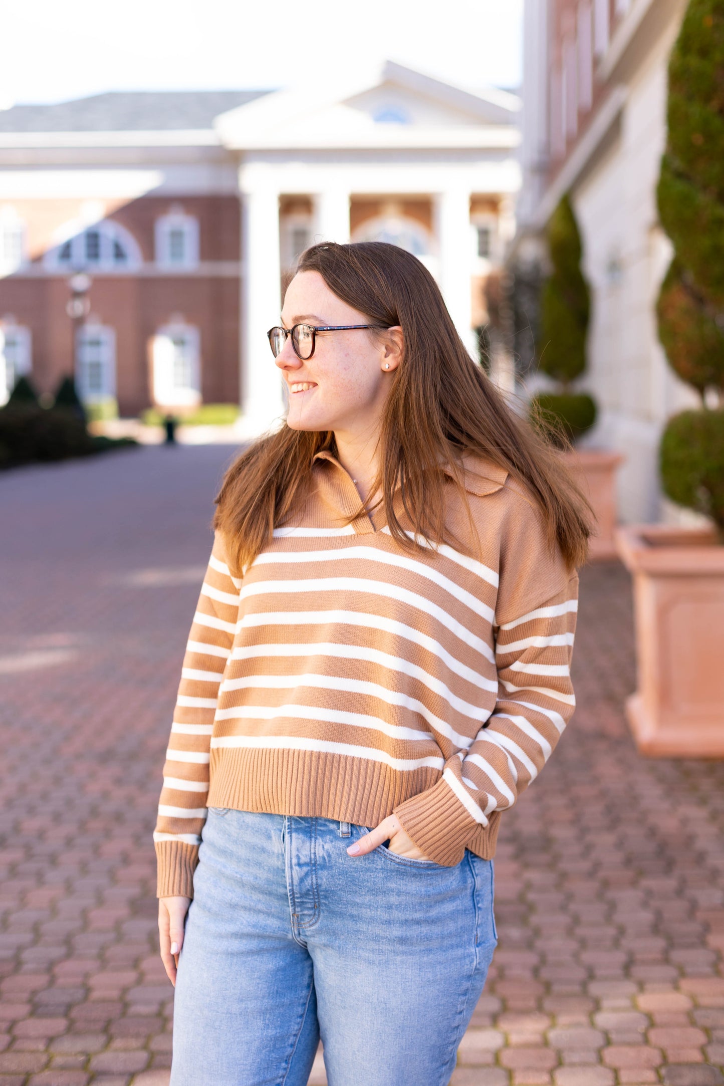 A woman in glasses confidently smiles outdoors near a brick building, wearing the chic Elliot Striped Sweater by Dogwood Cloth. Its viscose-polyester-nylon blend offers a stylish, modern cropped fit.