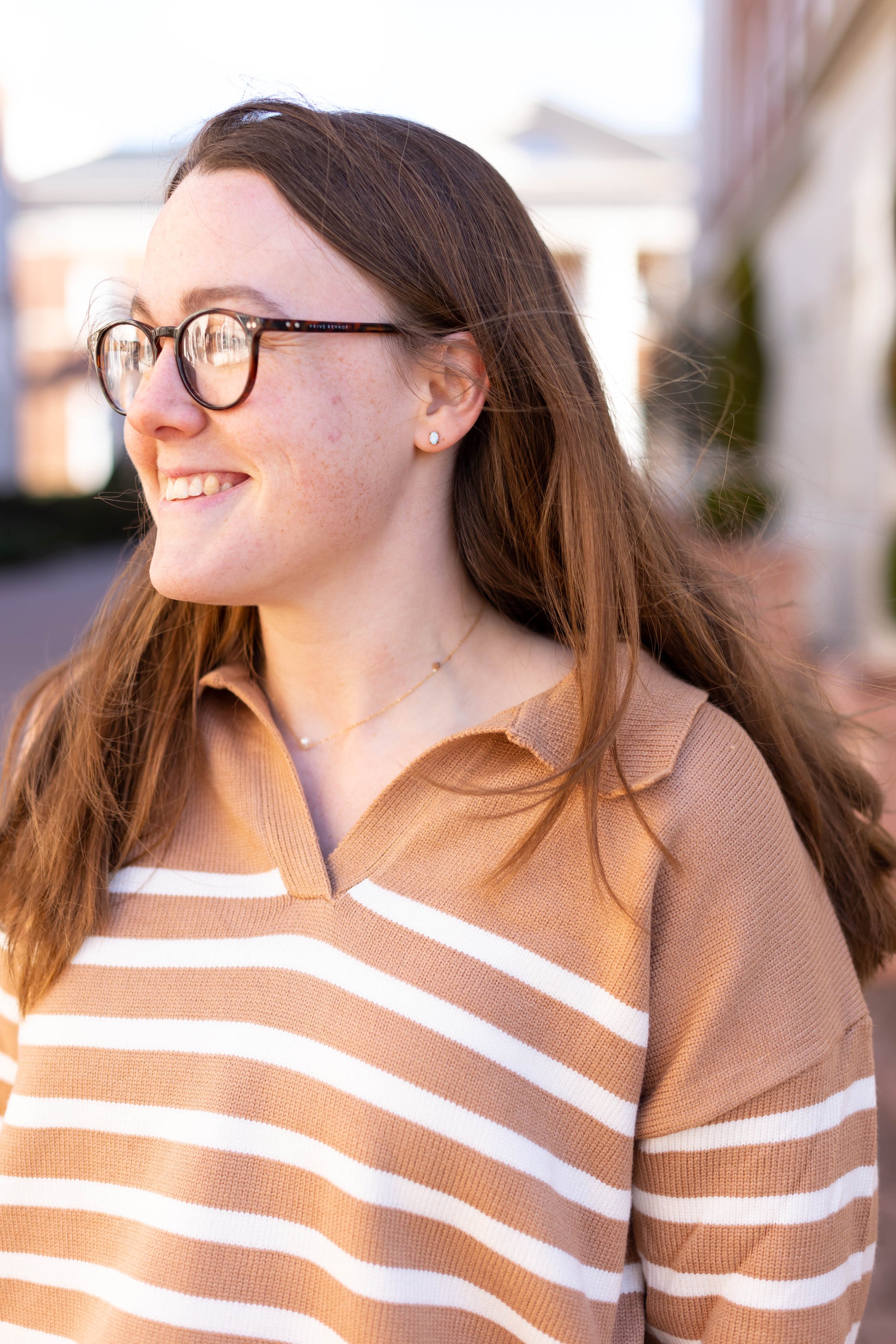 A long-haired woman with glasses is wearing the Dogwood Cloth Elliot Striped Sweater with a V-neckline. Smiling, she looks to the side, enjoying the outdoors.