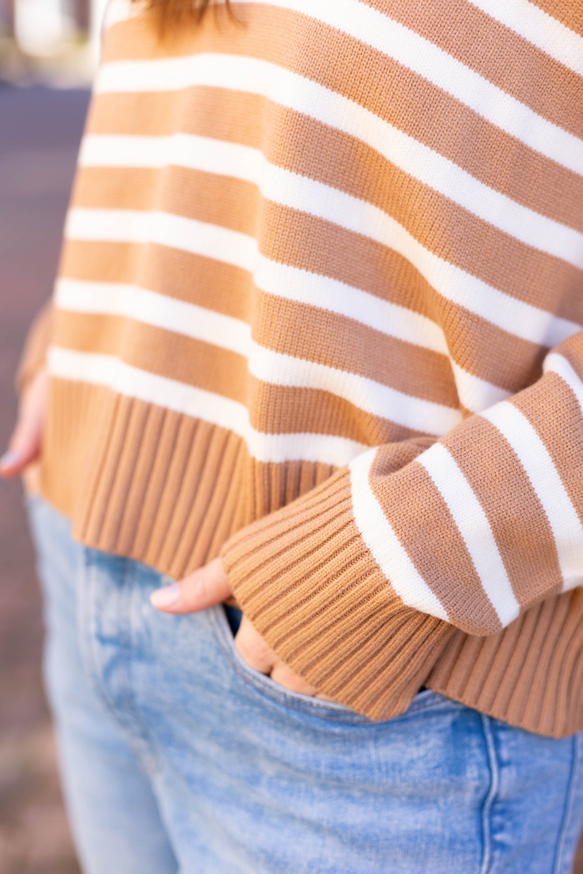 Someone is wearing the Elliot Striped Sweater by Dogwood Cloth, featuring brown and white stripes with a V-neckline, hands tucked into the pockets of light blue jeans.