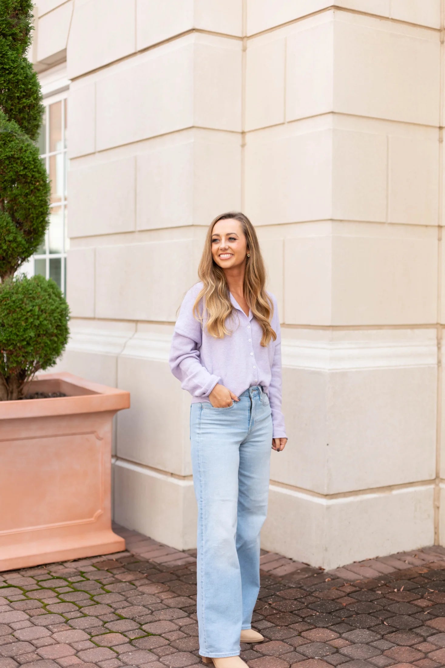 A person in a Dogwood Cloth Iris Lavender Sweater and blue jeans stands on a brick path, smiling warmly. The cozy knit fabric complements the large potted plant and the beige building in the background, creating a harmonious scene.