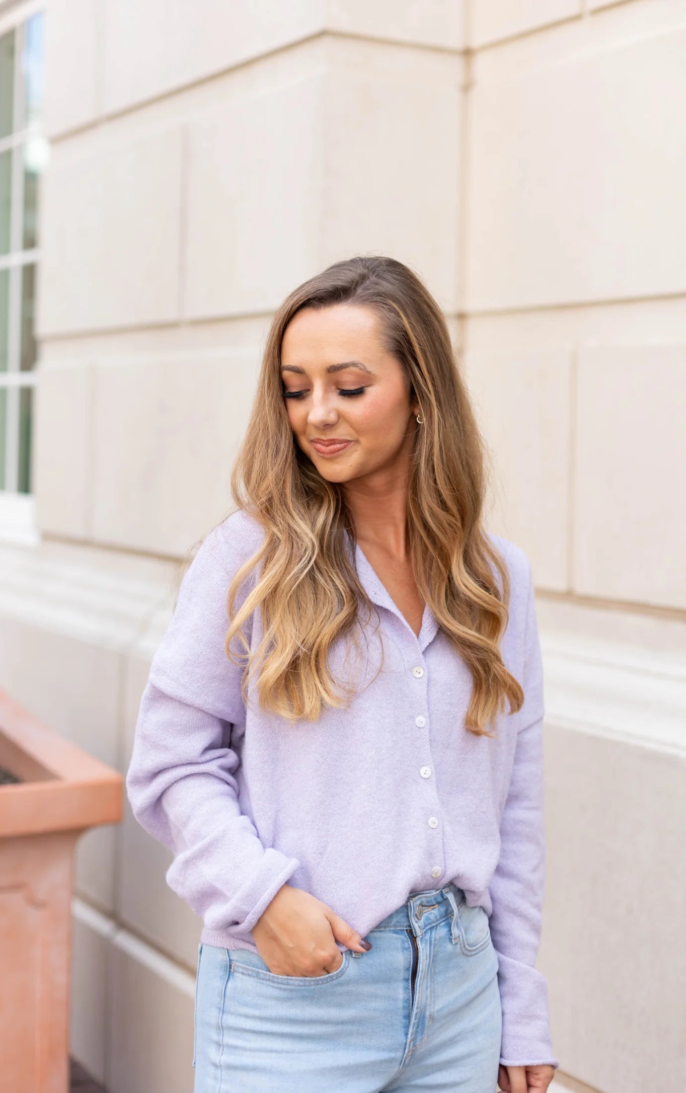 A person with long hair is standing near a light-colored wall, wearing the Dogwood Cloth Iris Lavender Sweater and jeans, looking down.