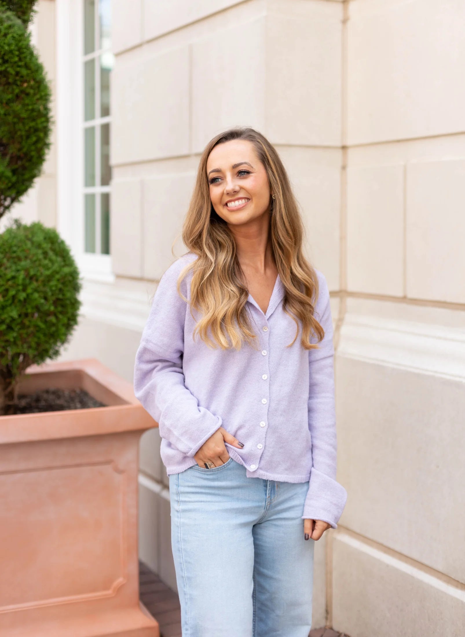 A woman in a Dogwood Cloth Iris Lavender Sweater and jeans stands smiling in front of a beige building with a potted plant.