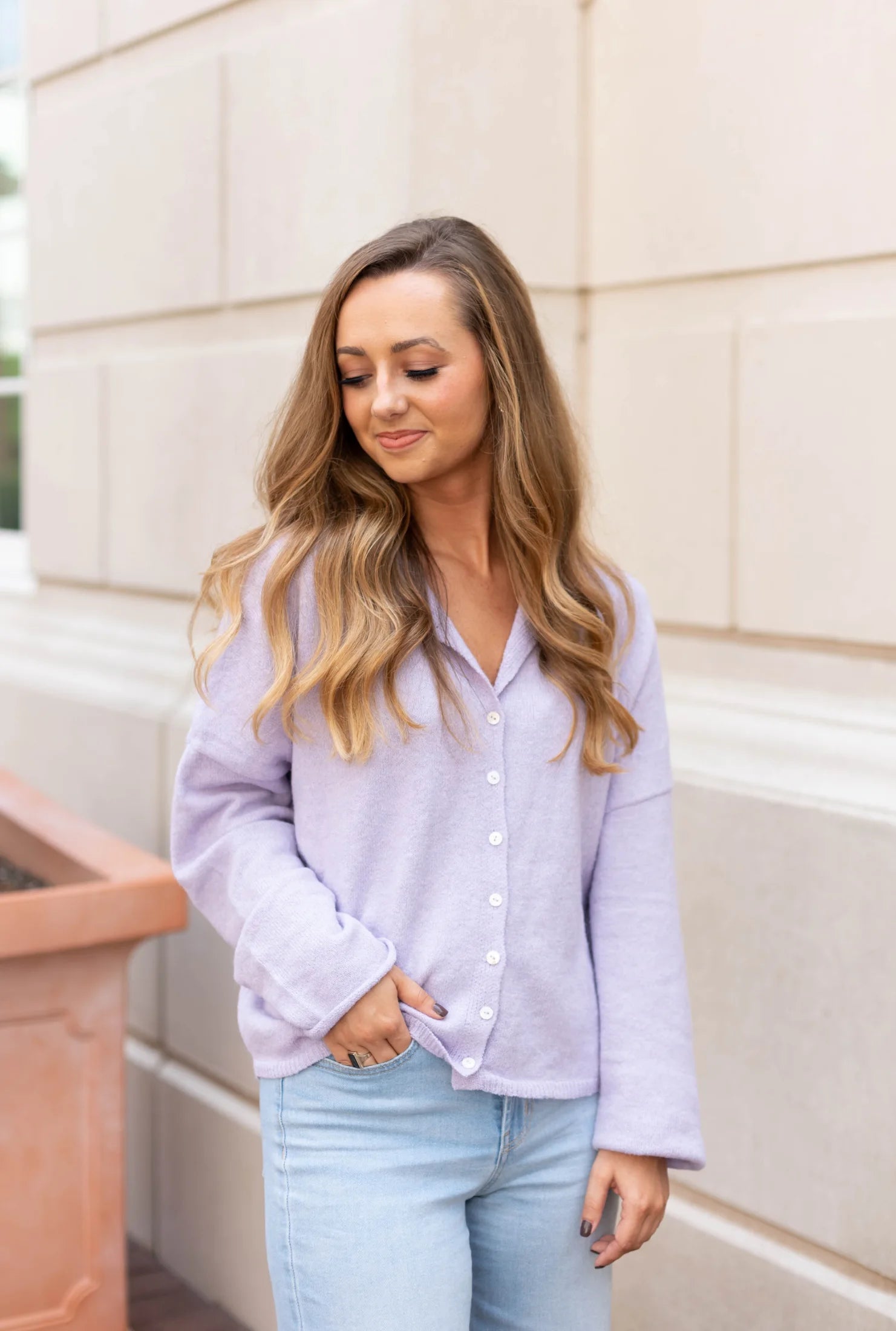 A woman dressed in the Iris Lavender Sweater from Dogwood Cloth and jeans stands with one hand in her pocket against a light-colored wall, gazing slightly downward.