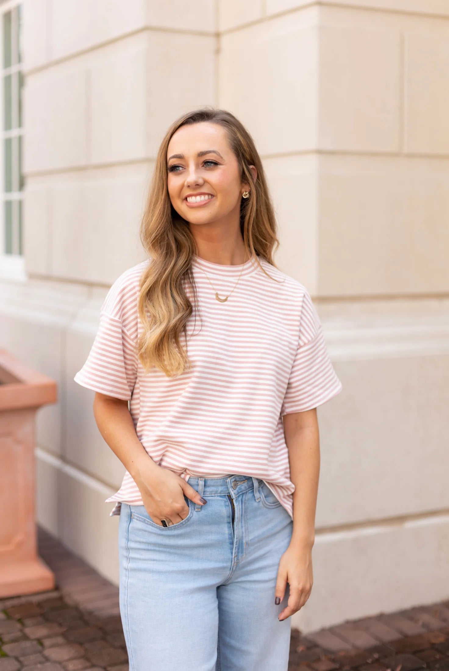 A person with long hair smiles, dressed in the Delaney Striped Tee from Dogwood Cloth and jeans, radiating casual comfort while standing in front of a beige building.