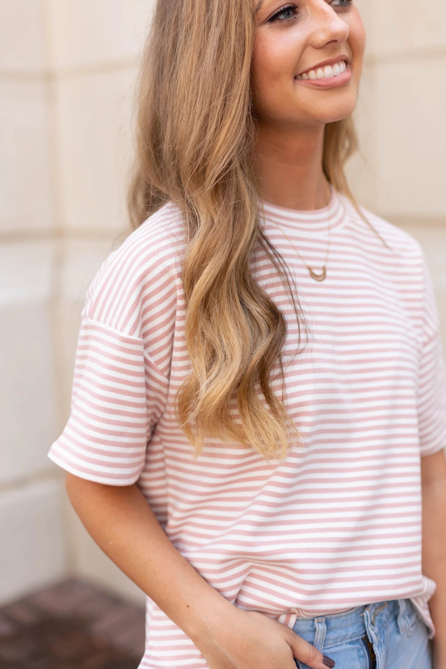 A person wearing a Dogwood Cloth Delaney Striped Tee and jeans, with long wavy hair, stands and smiles, exuding a sense of casual comfort.