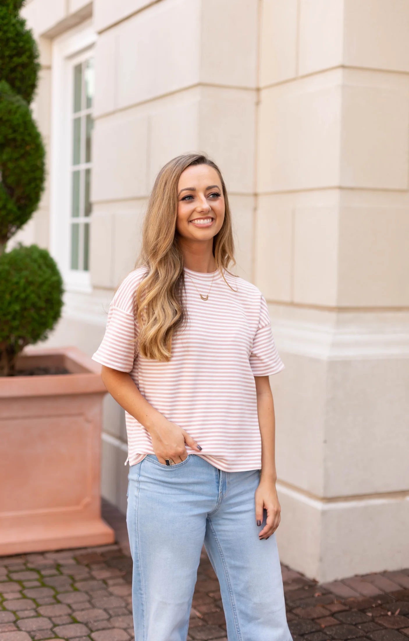 Wearing the versatile Delaney Striped Tee from Dogwood Cloth paired with jeans, a woman stands smiling in front of a beige building with a potted plant, exuding casual comfort.